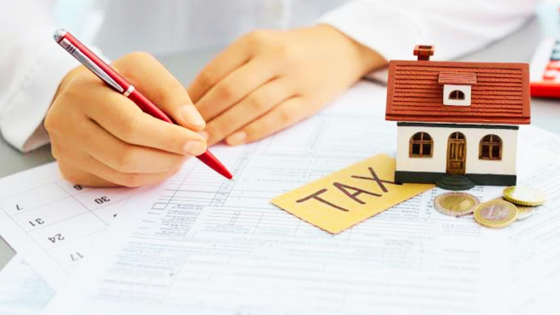 Hand with red pen over tax forms beside miniature house, yellow TAX tag, and coins on a desk.