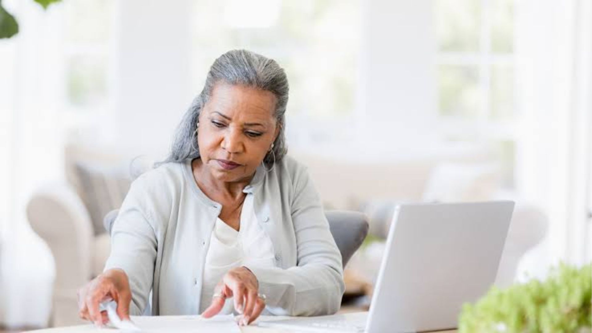 Older woman with gray hair in light cardigan reviewing documents at table with laptop in bright home office.
