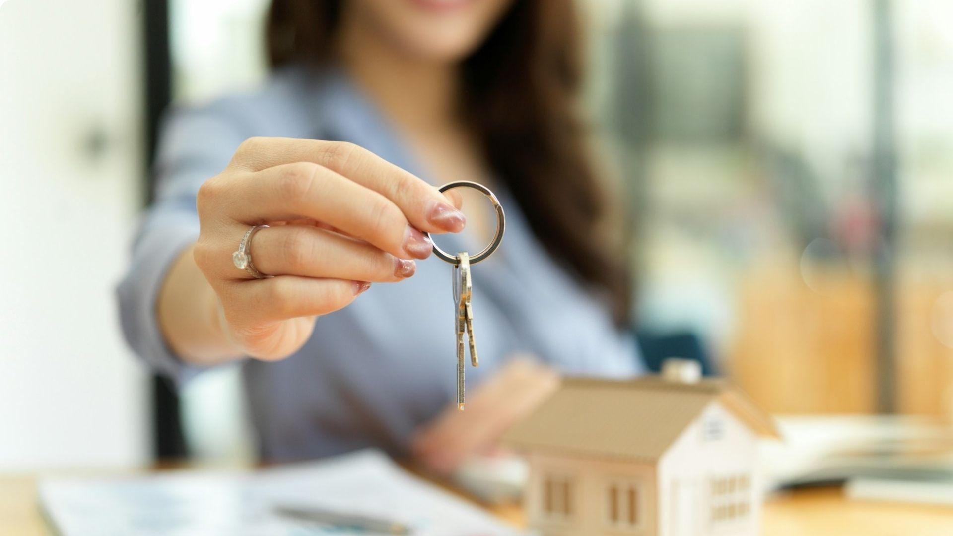 Woman offering house keys with small model house on desk in real estate closing