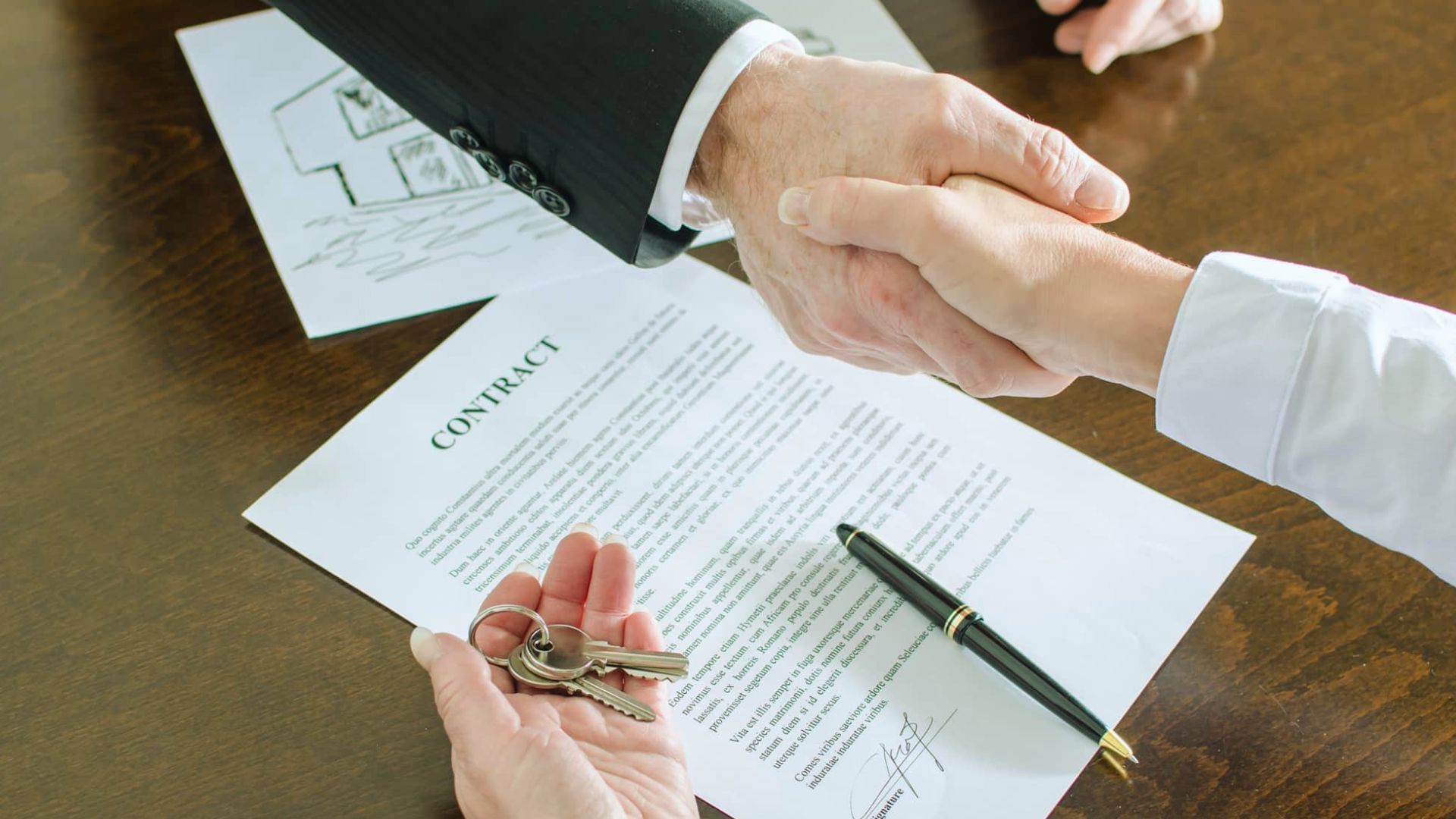 Two people shaking hands over signed property contract while holding house keys on table