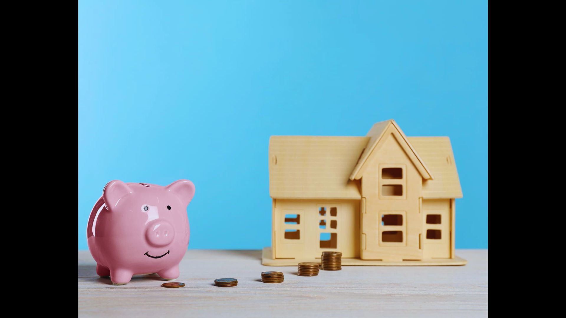 Pink piggy bank facing a wooden model house with a path of small coins between them on a light blue surface.