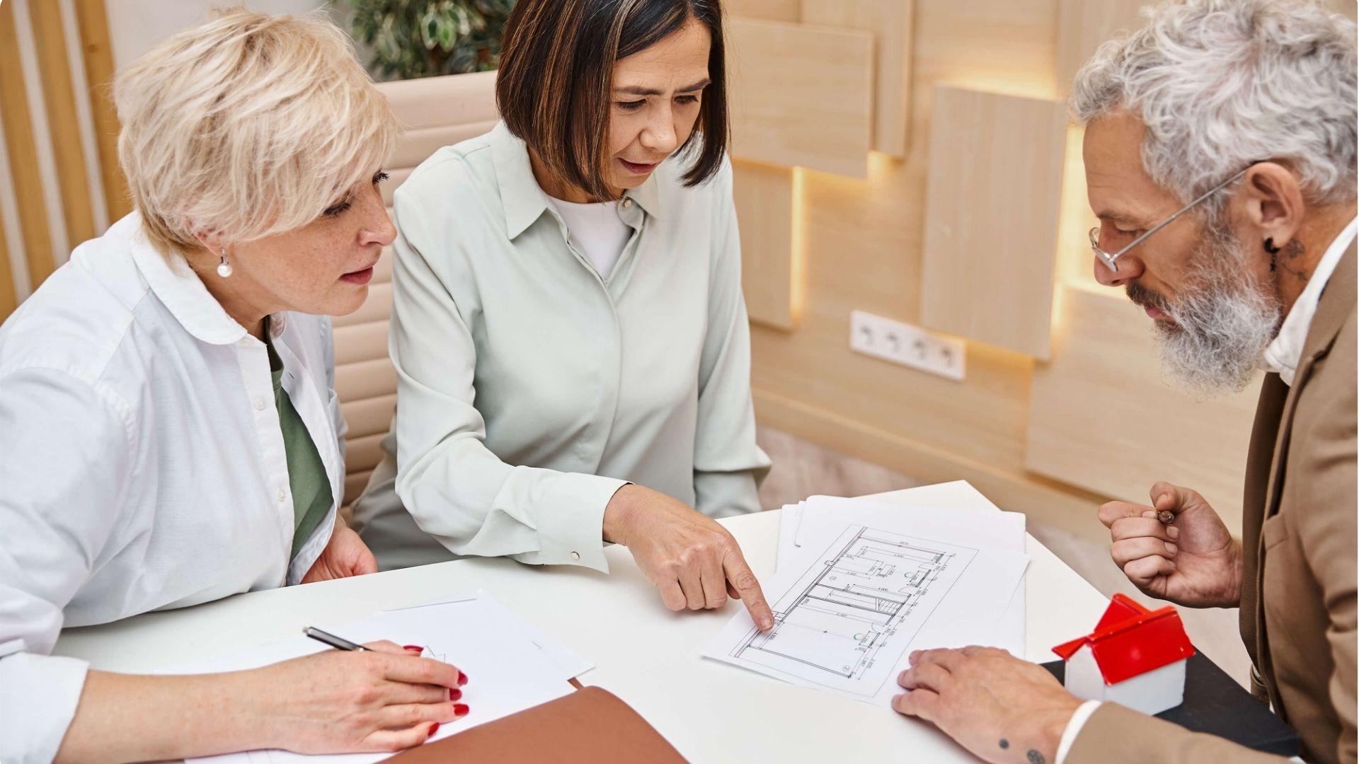 Two women and bearded man reviewing house papers at table with red-roof model home