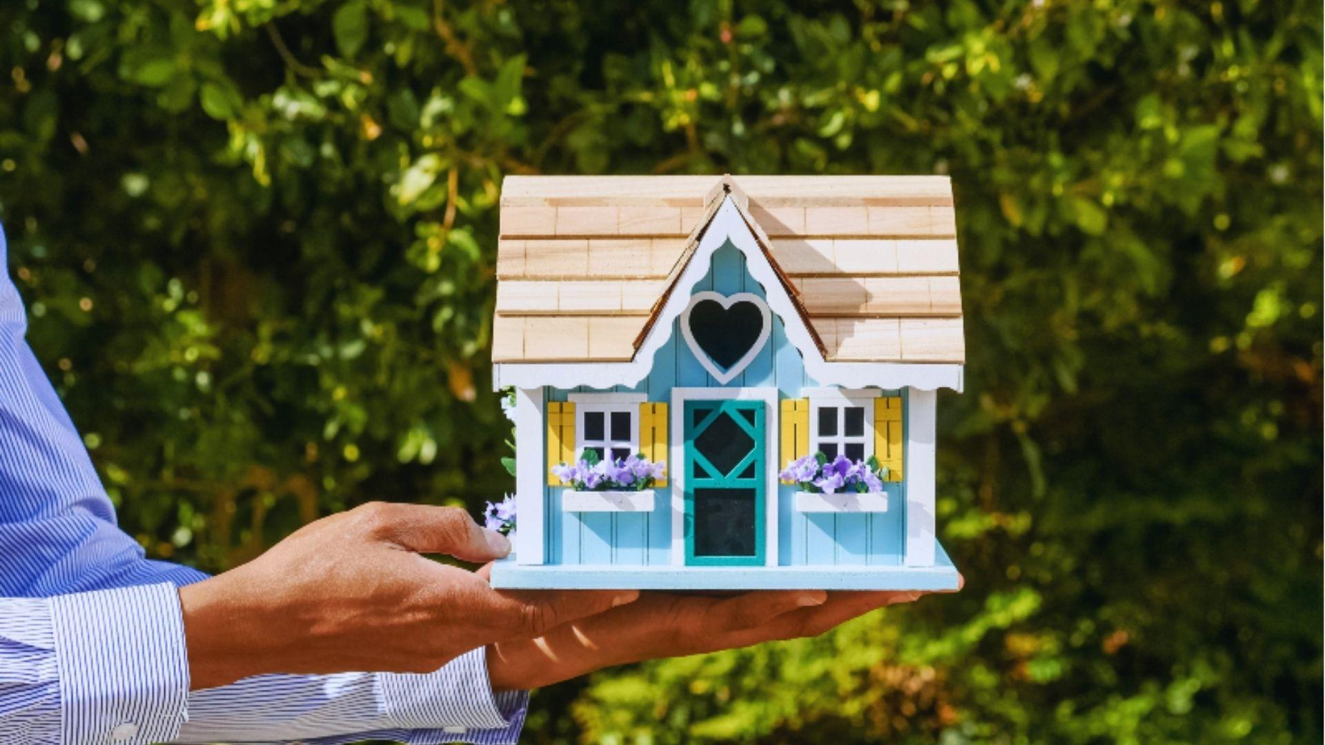 Person's hands in a blue striped shirt holding a small decorative blue model house with light wood roof