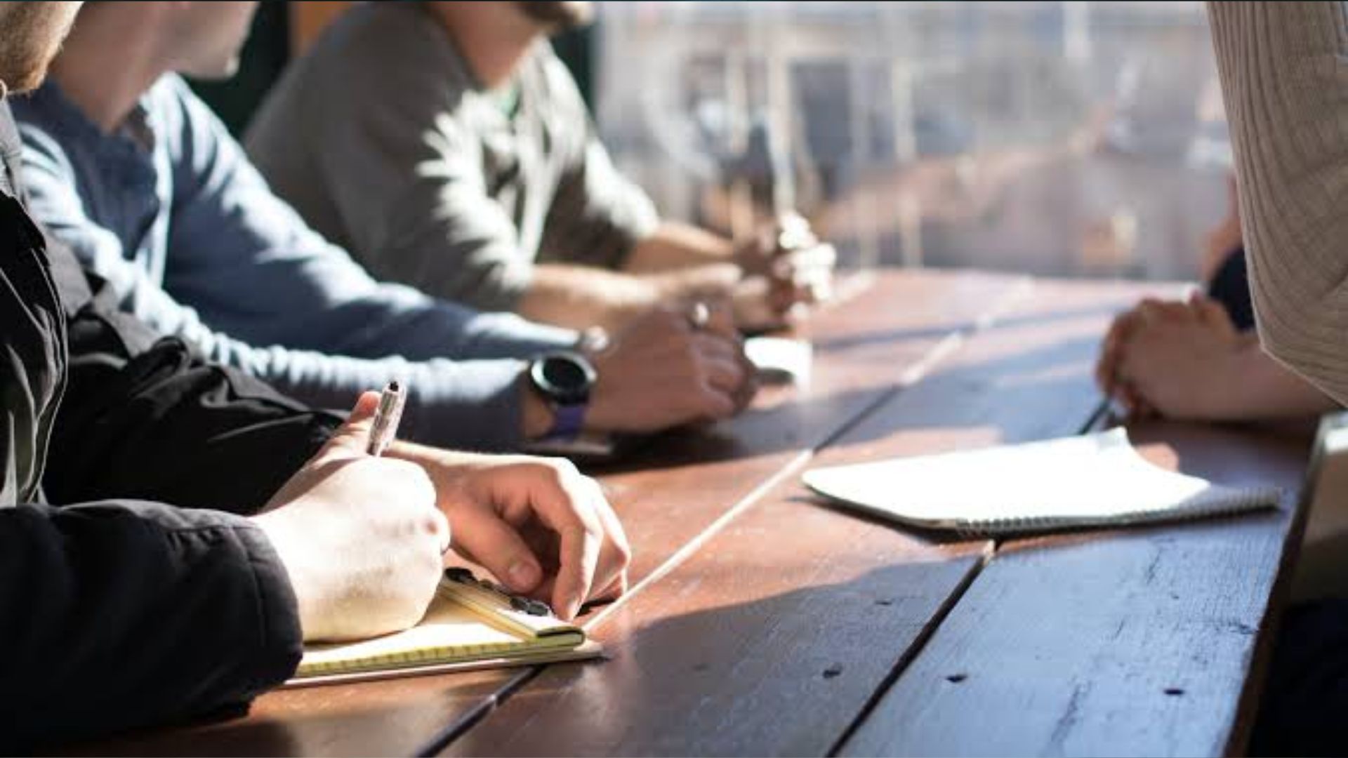Close-up of people taking notes at a long wooden table during a sunny meeting, hands holding pens and notebooks.