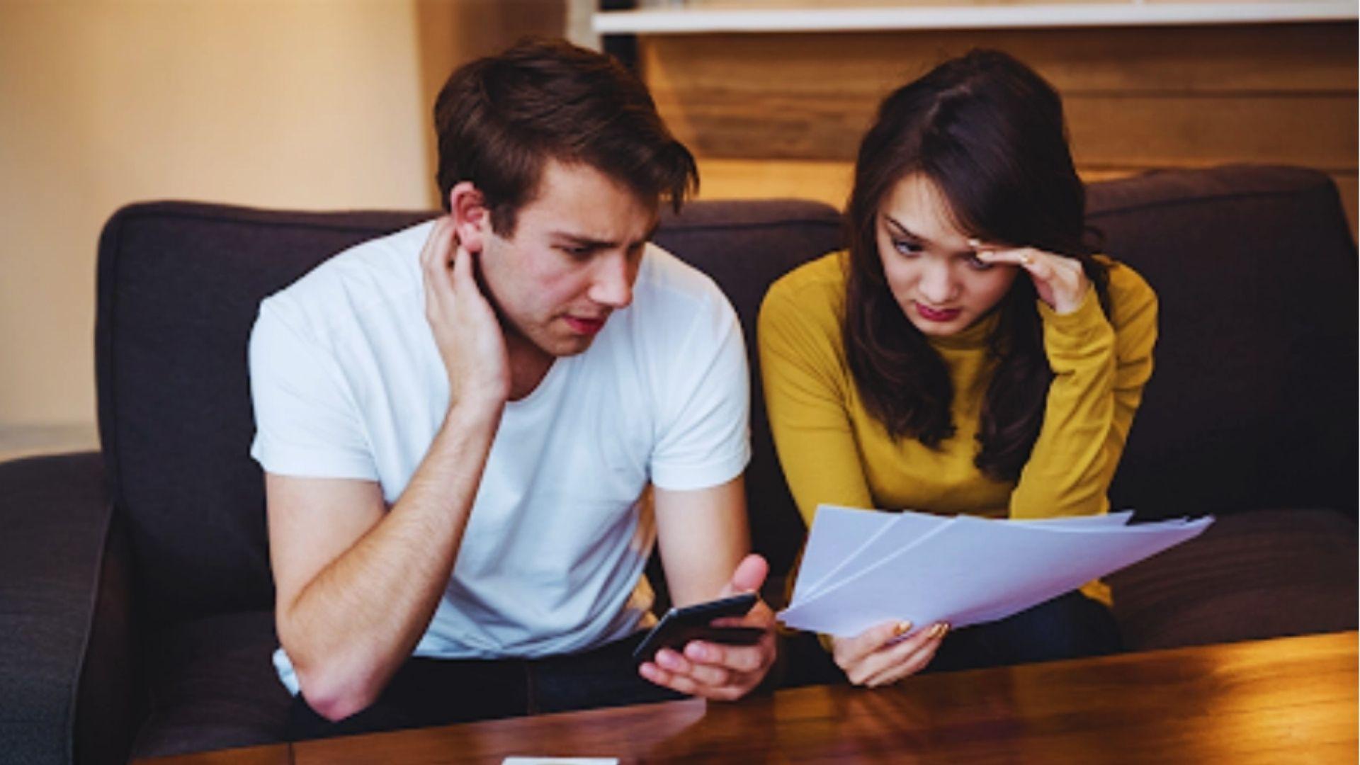 Stressed young couple on a couch reviewing papers and phone, man touching neck, woman holding bills with hand on forehead.