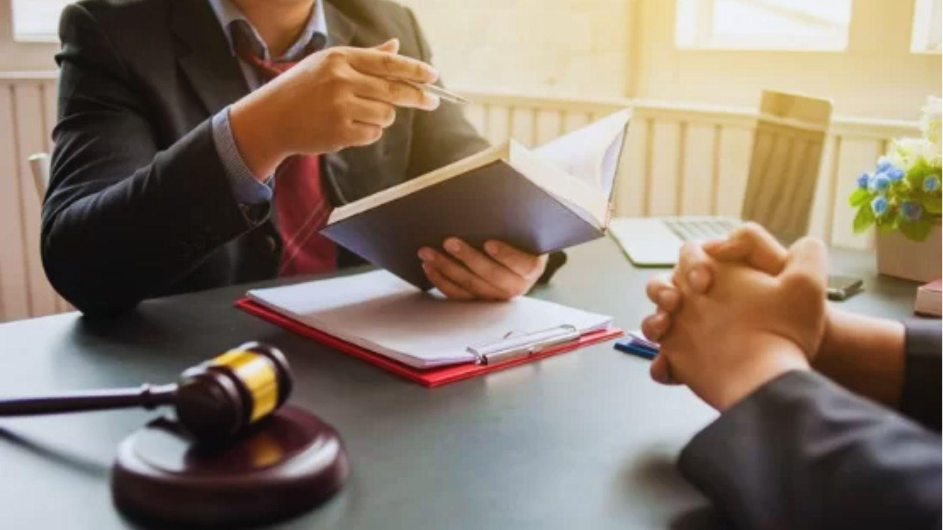Lawyer in a dark suit holding a pen and an open book while explaining documents on a red clipboard across a desk