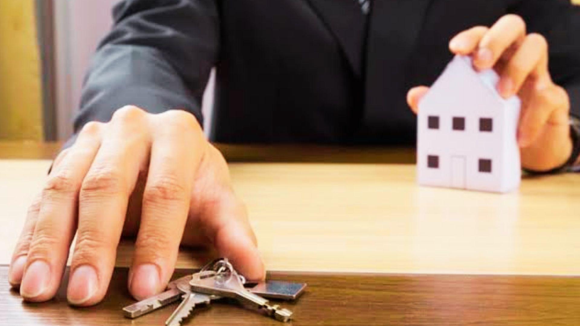 A person holding a small white paper house model while reaching for a set of keys on a wooden desk.