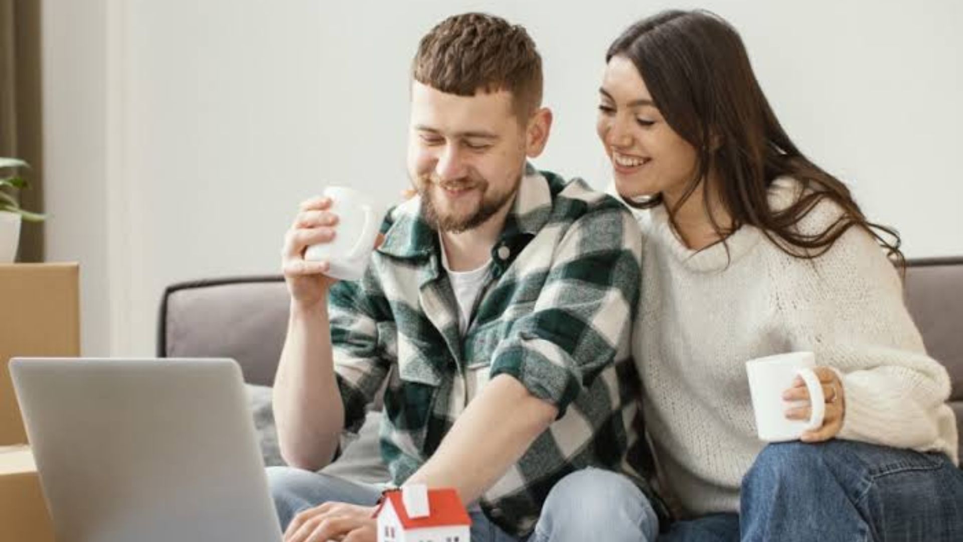 Happy couple holding white mugs sitting on couch looking at laptop with small red roof model house on table.