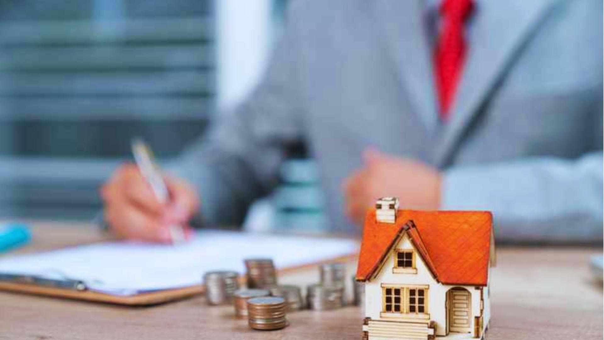 Small model house with stacks of coins on a desk, person in suit signing paperwork in blurred background.