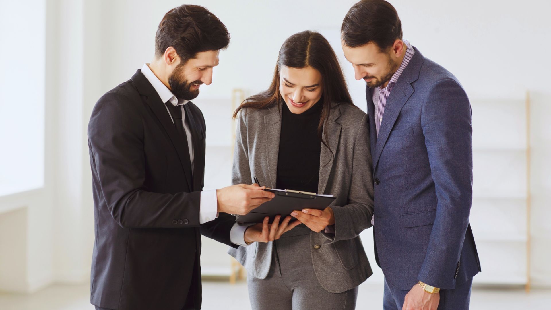 Three business professionals smiling together while looking at a clipboard held in a bright, modern office.