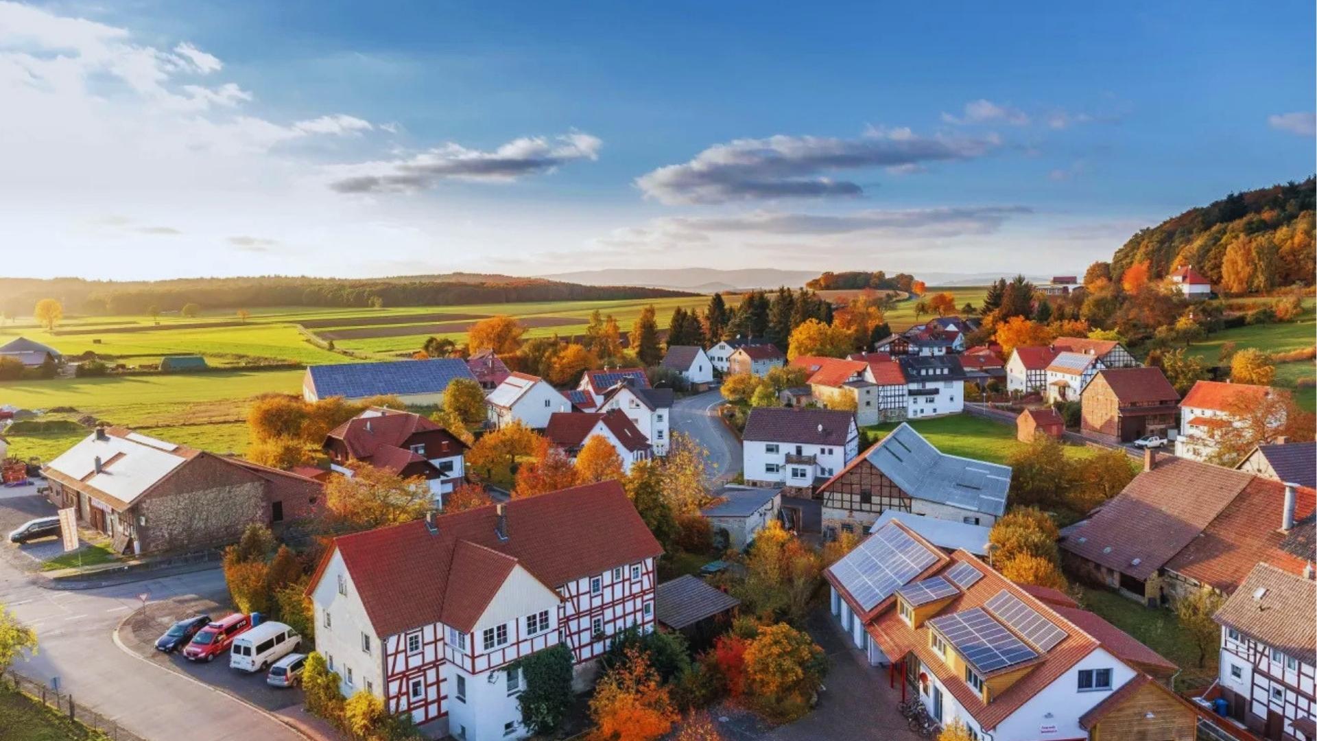 Aerial view of a quaint village with red roofs surrounded by green fields and autumn trees.
