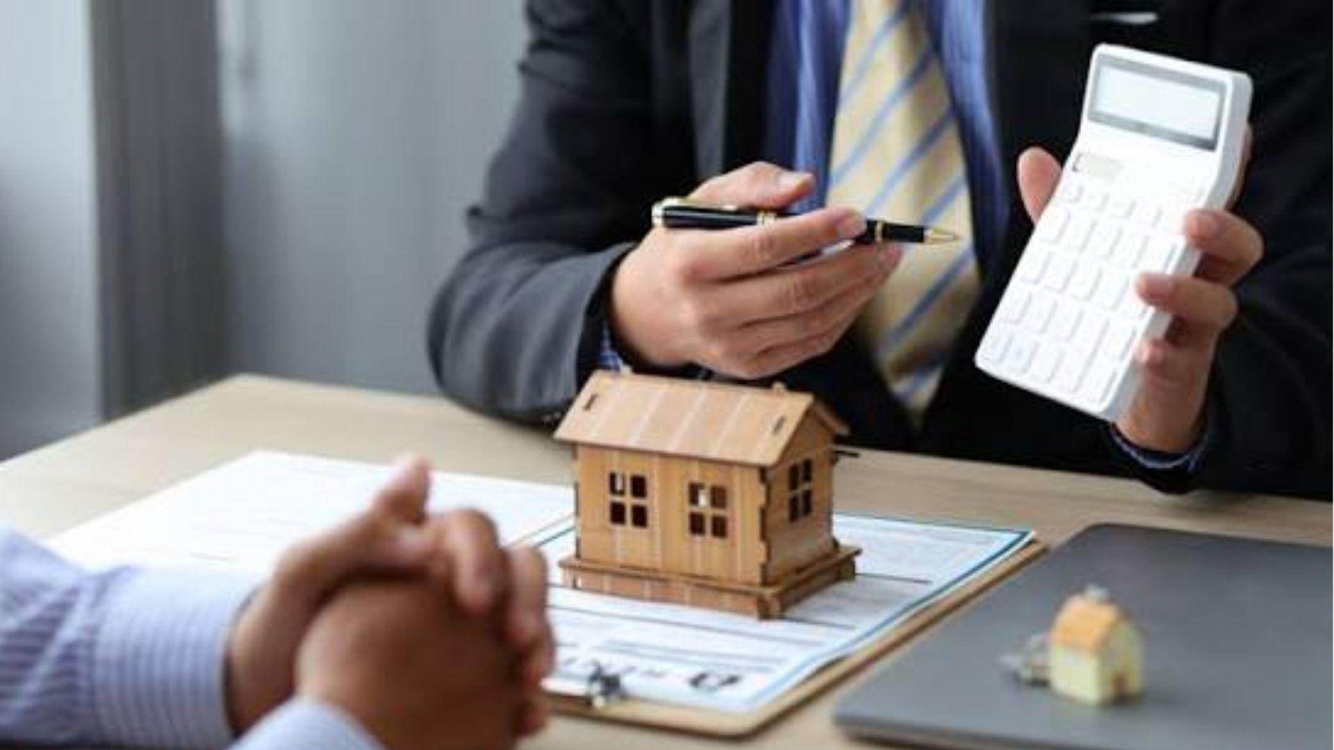 Person in suit holding a black pen and white calculator over a small wooden model house on paperwork, with another person's clasped hands visible in foreground.