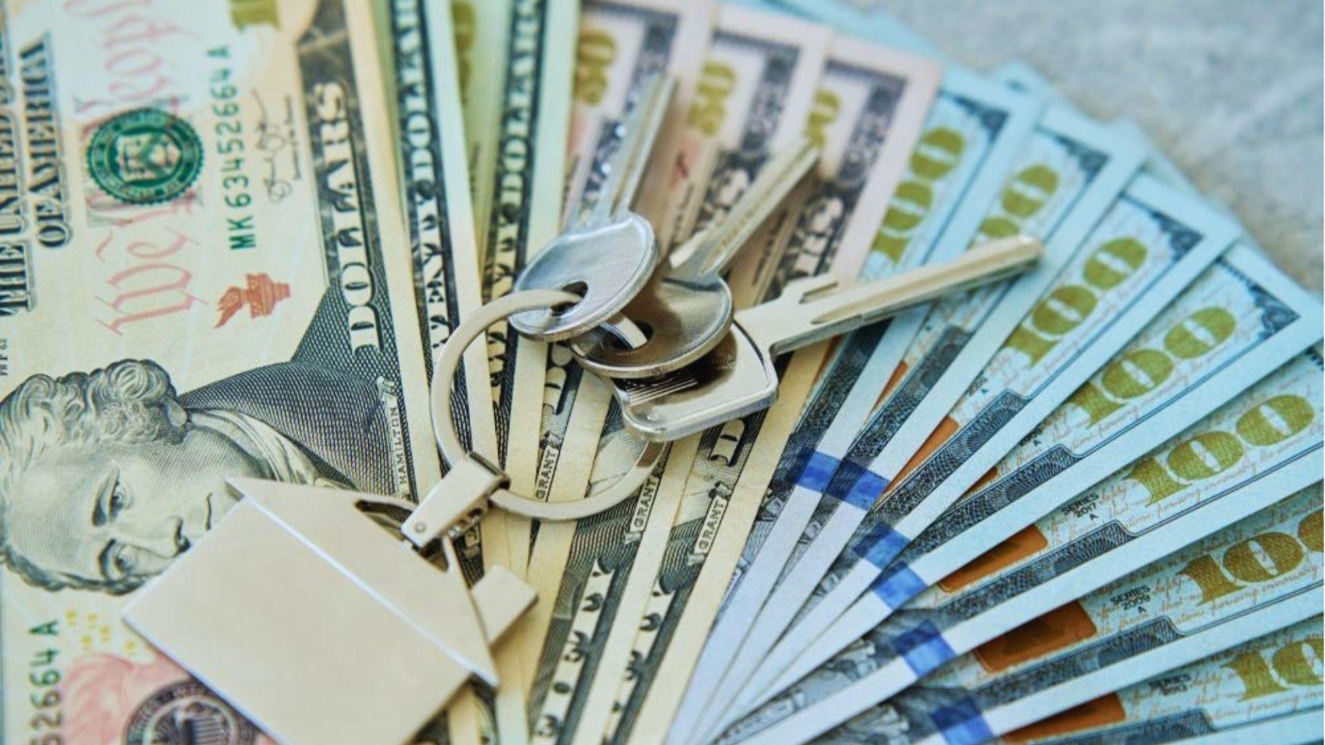 House keys with home-shaped keychain resting on fanned US hundred and ten dollar bills.