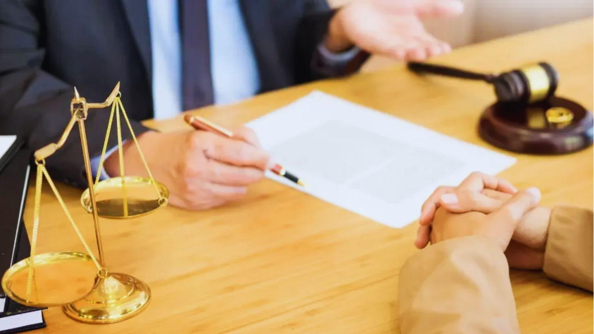 Lawyer in suit holding pen over document and gesturing across wooden desk to client with clasped hands, gold scales of justice in foreground and gavel on stand in background.
