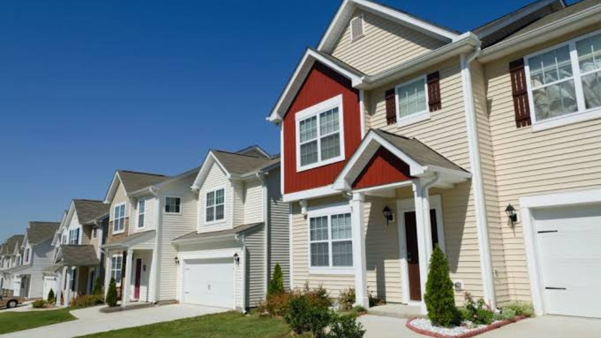 Row of beige two-story townhouses with red gable accents, white garage doors, small shrubs and lawns under clear bright blue sky.