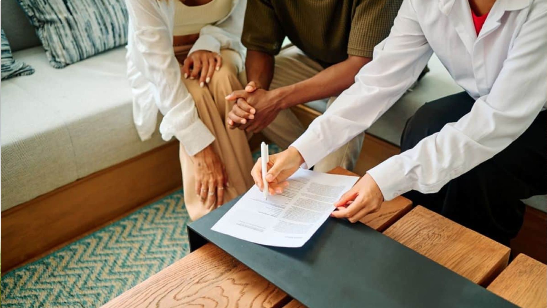 Professional in white shirt pointing at contract with pen on low table, couple with clasped hands watching from couch, consultation scene.