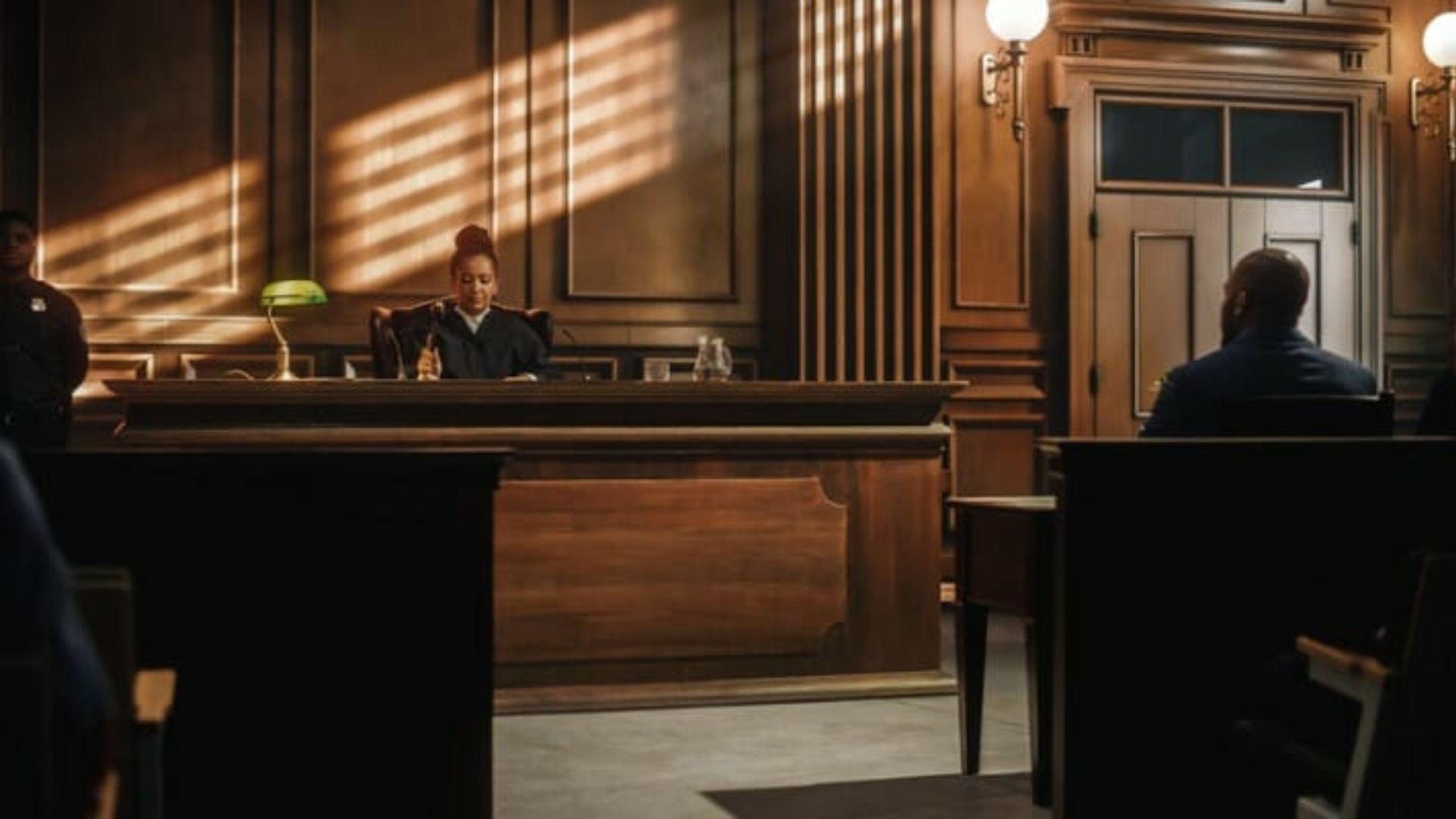 Female judge holding gavel at wood-paneled bench in sunlit courtroom with blind shadows, man seated at table viewed from behind.