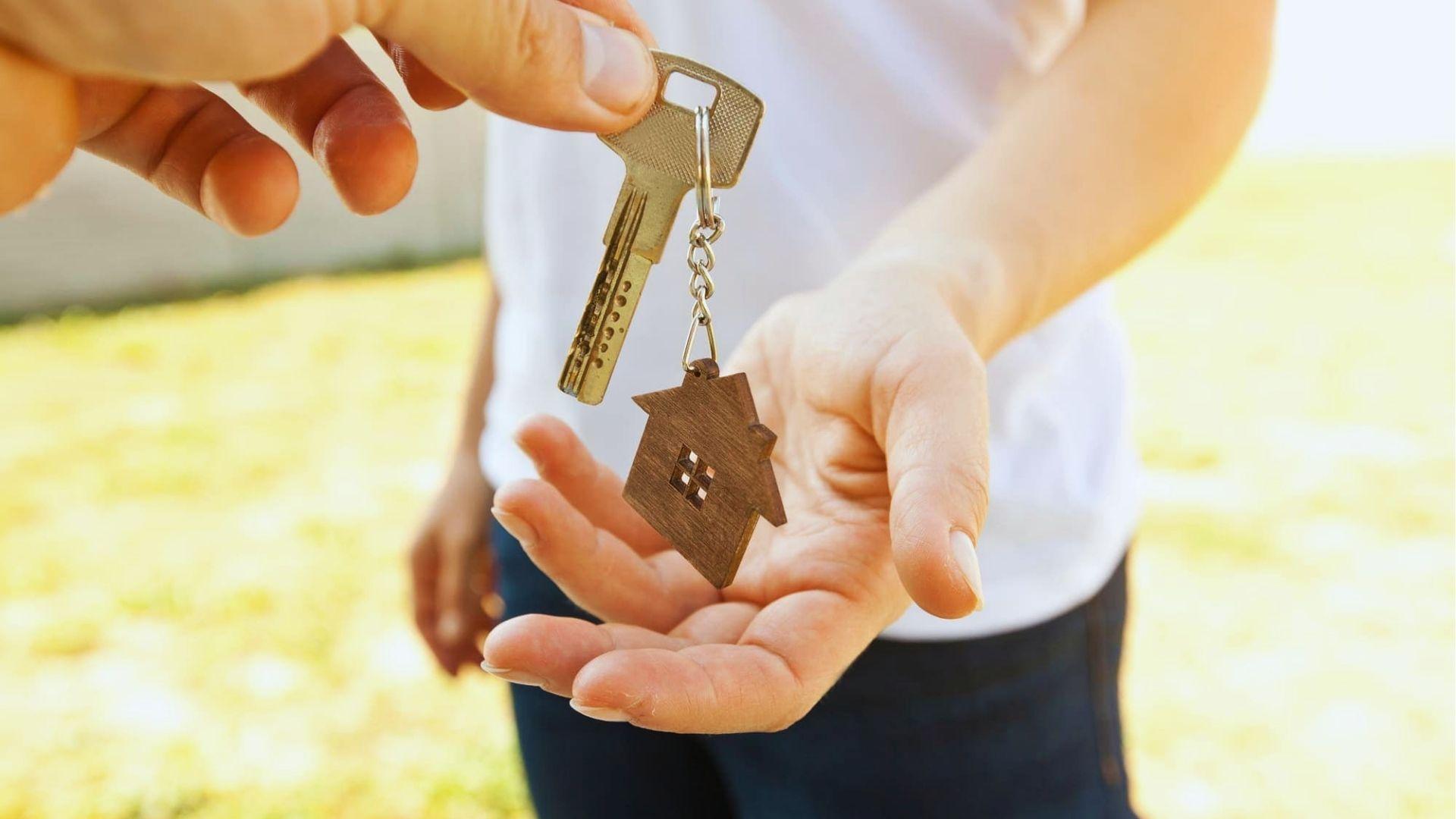 Close-up of one hand passing a brass house key with a small wooden house-shaped keychain.