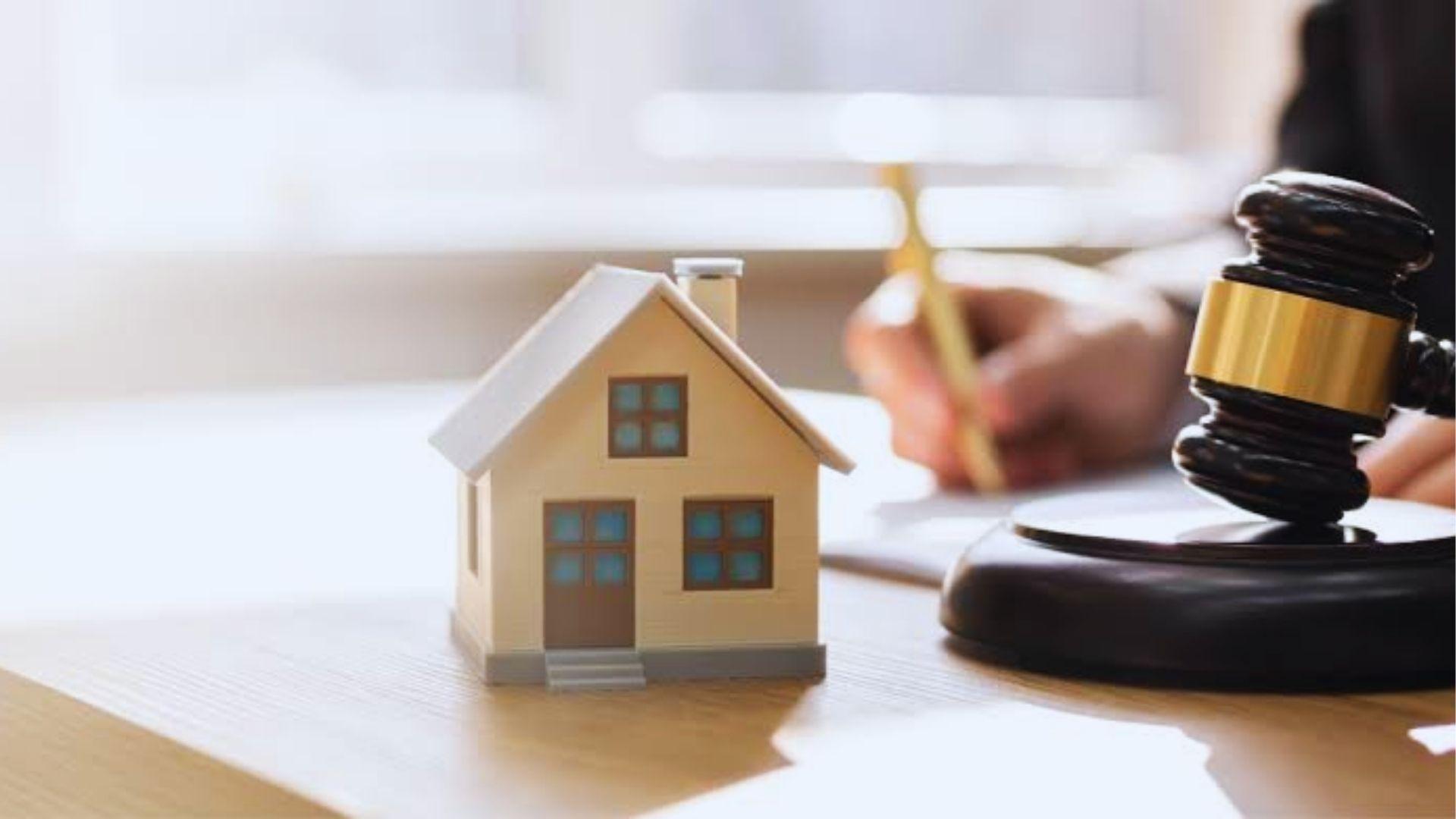 Small beige model house with brown door and blue windows sitting on a desk next to a wooden judge's gavel and sounding block
