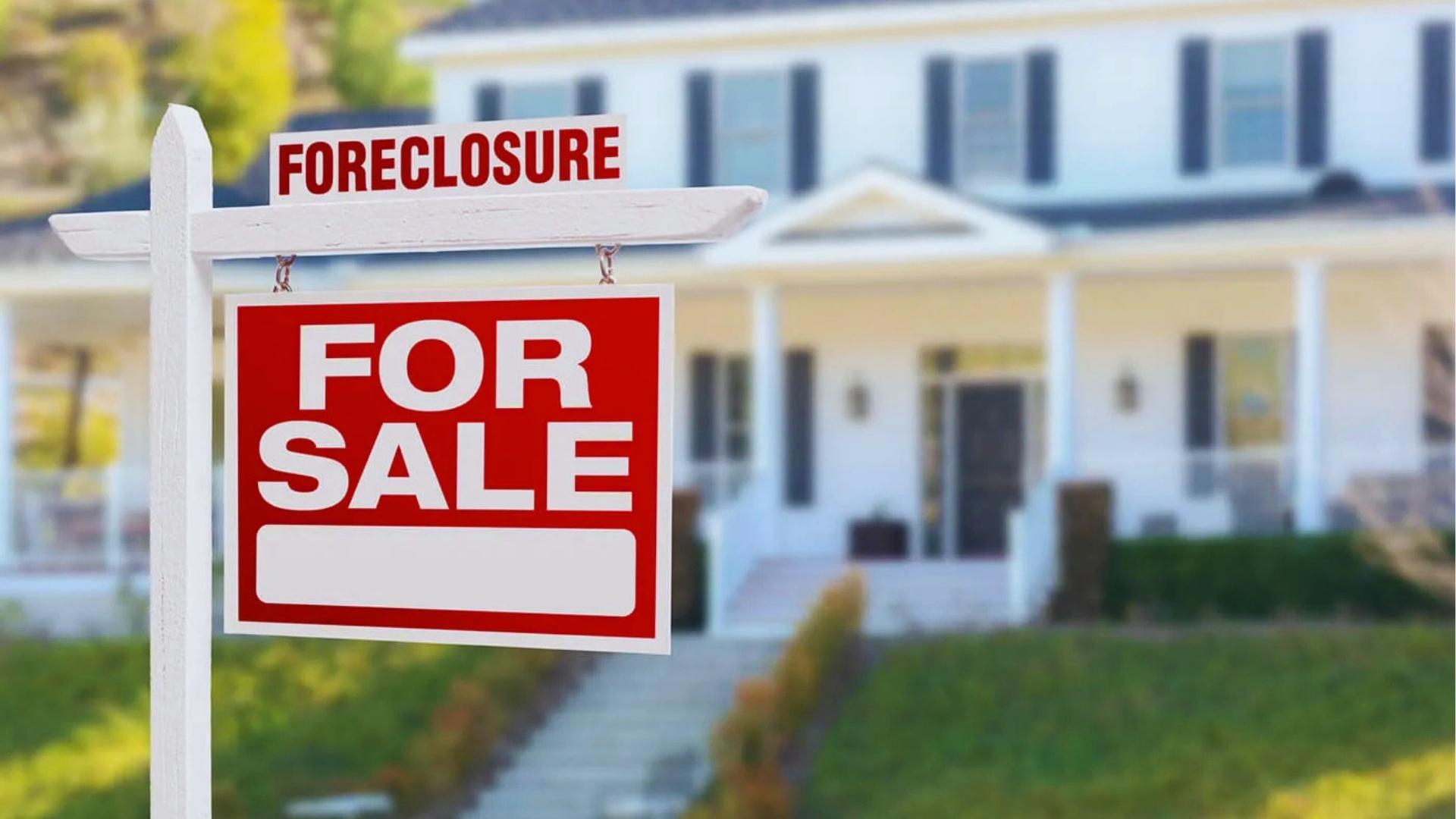 Red foreclosure for sale sign standing in front of white suburban house with green lawn.