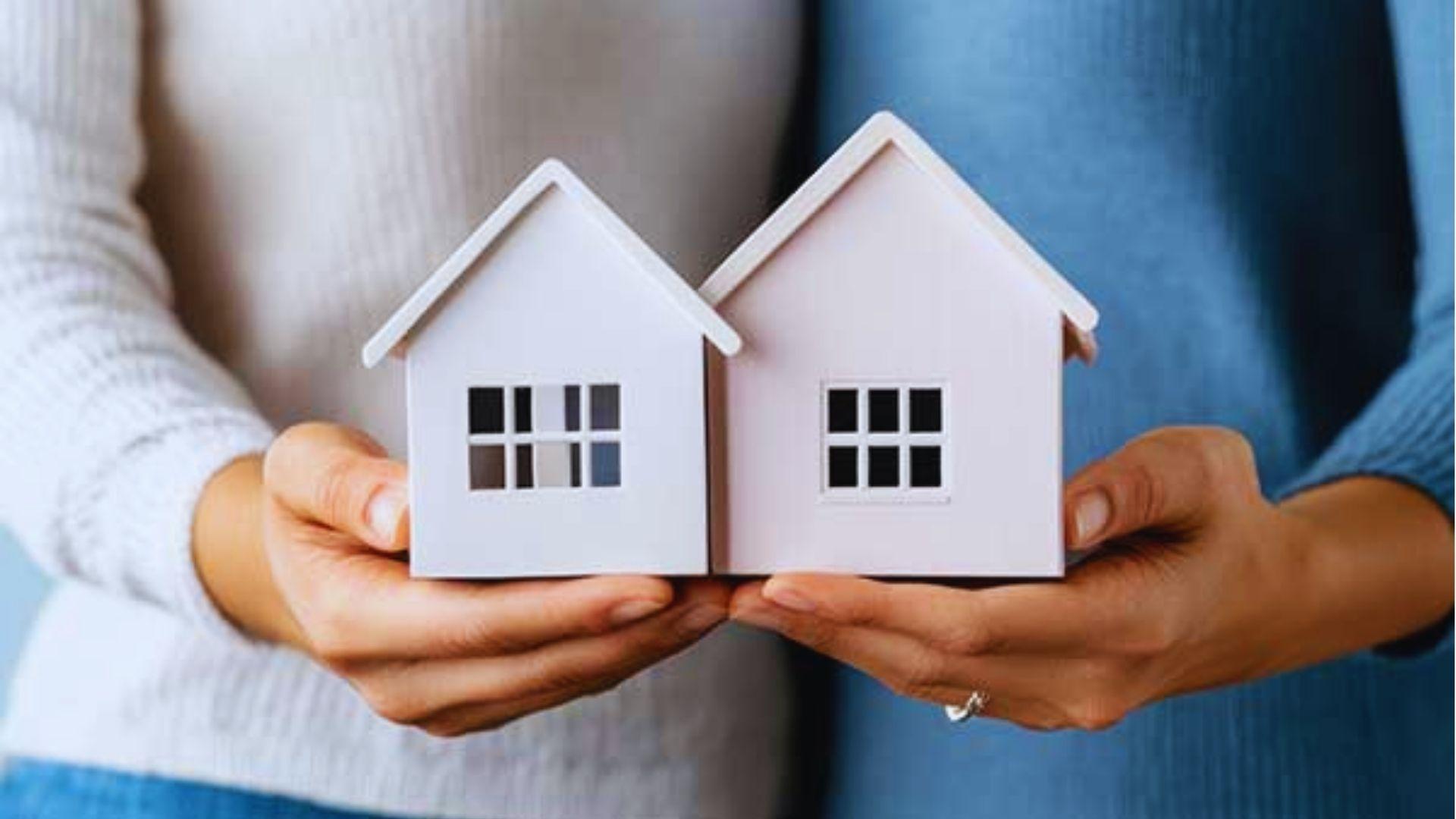 Two people holding two small white model houses side by side in open hands.