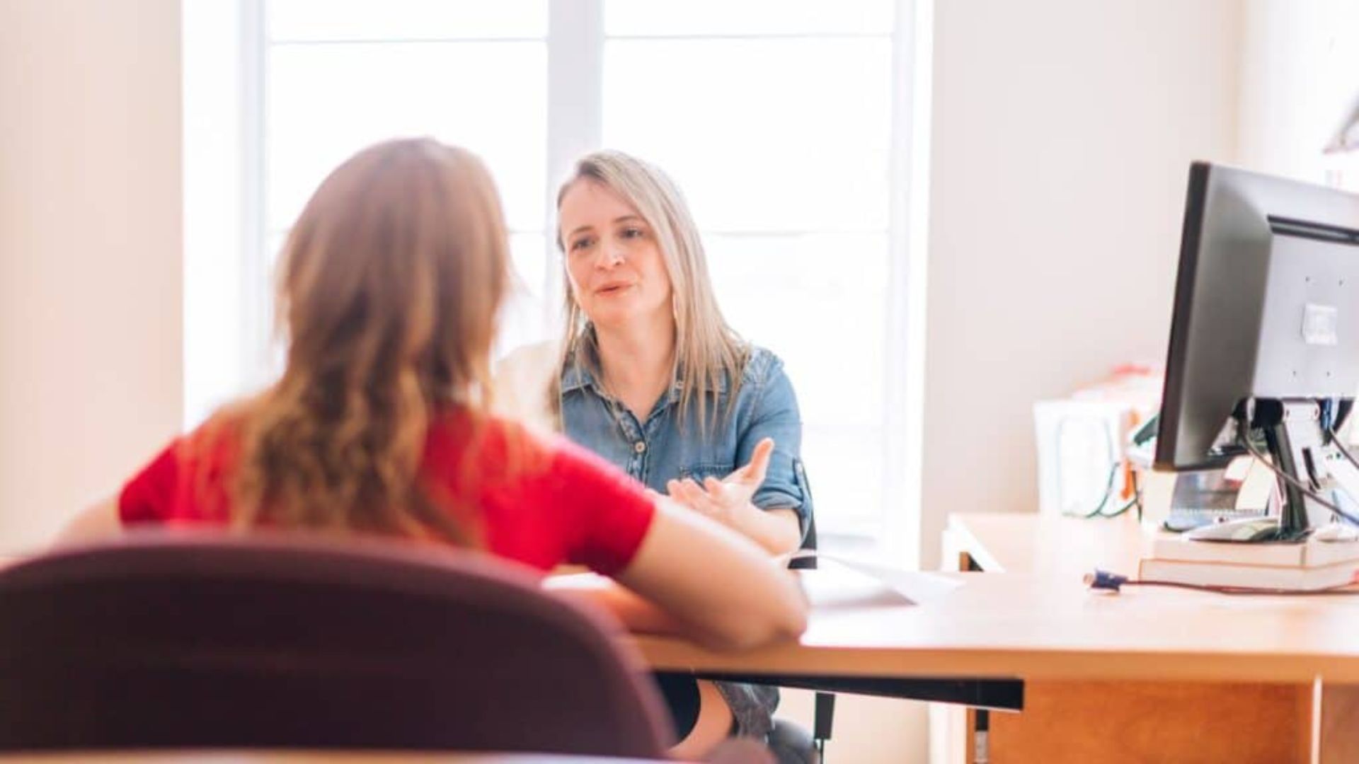Woman in denim shirt gesturing while counseling woman in red shirt across bright office desk with computer monitor.