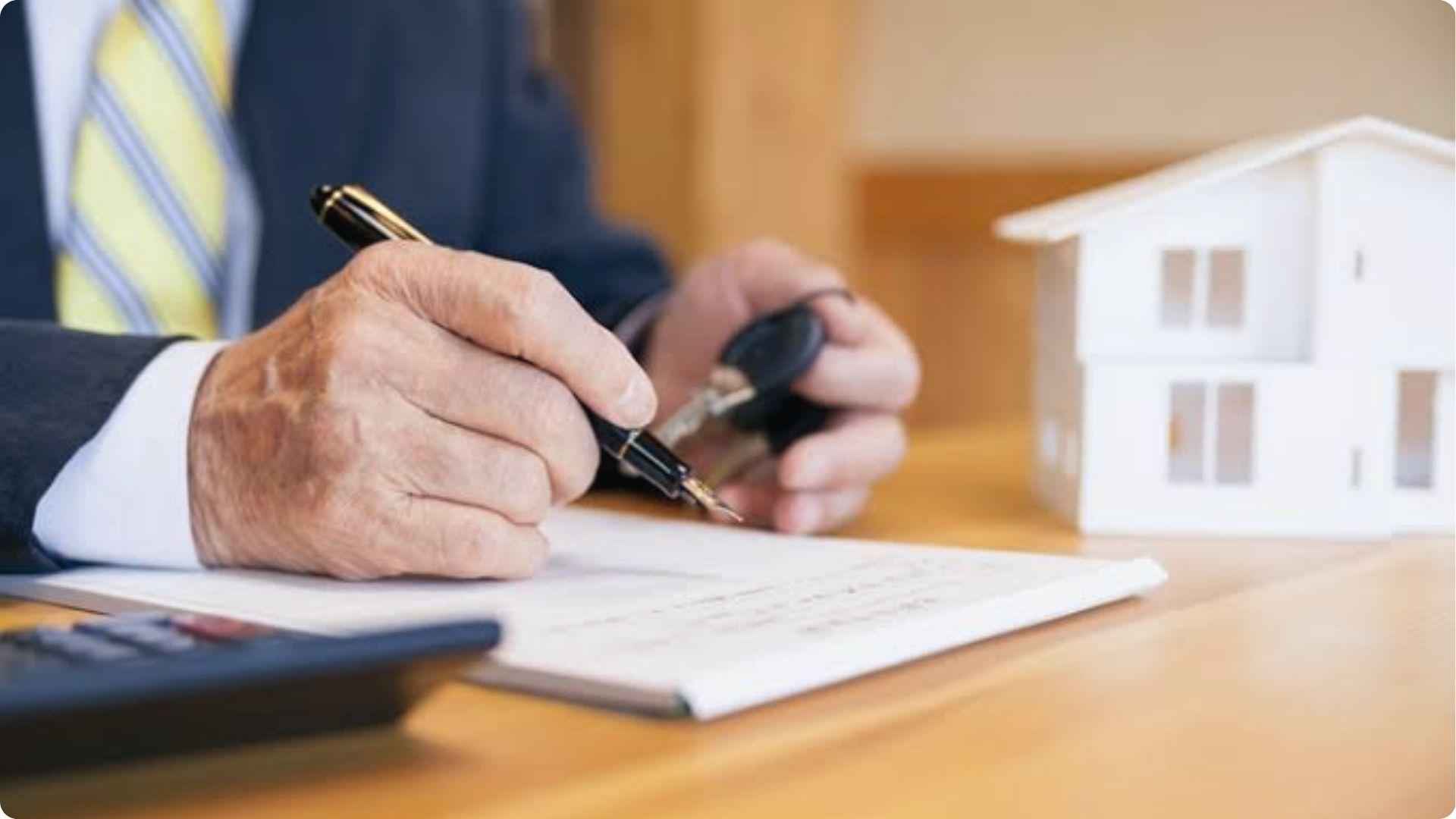 Person signing home loan documents while holding house keys beside white model house on table.