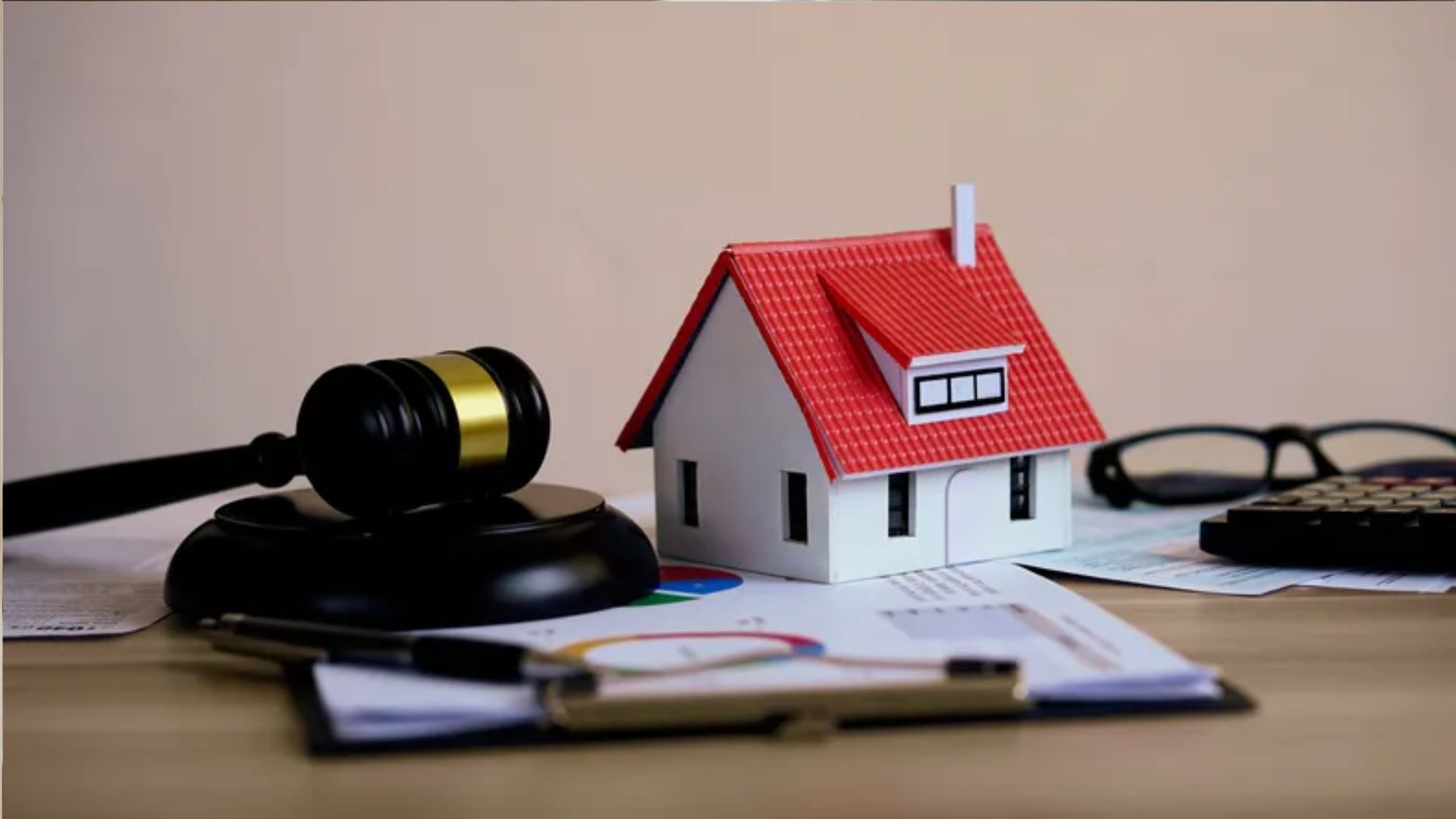 Small white house with red roof next to judge gavel on stand, with calculator, glasses and financial charts on desk.