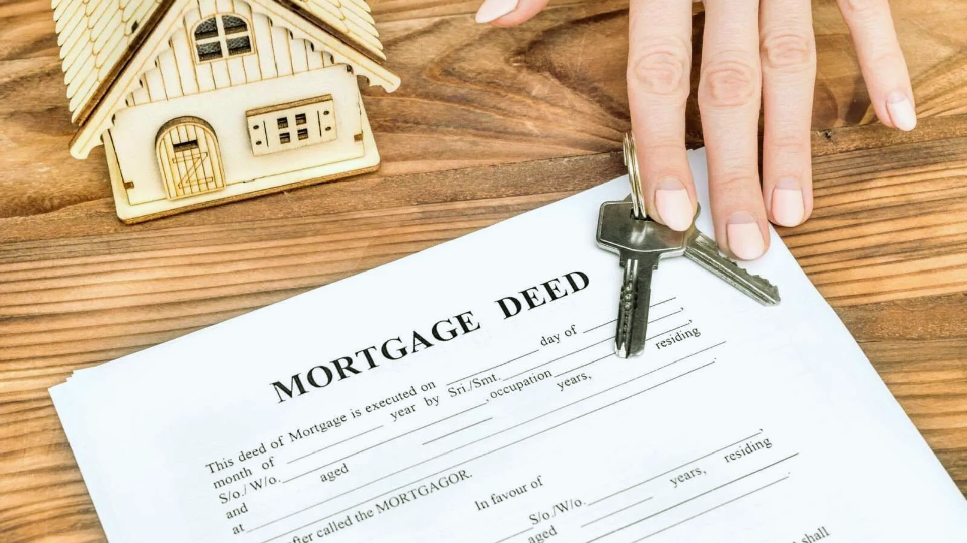 Mortgage deed document with wooden house model and hand holding house keys on wooden desk