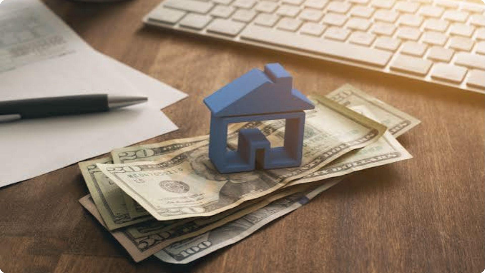 Small blue house model sitting on stacked US dollars on a desk with keyboard, pen and papers nearby.