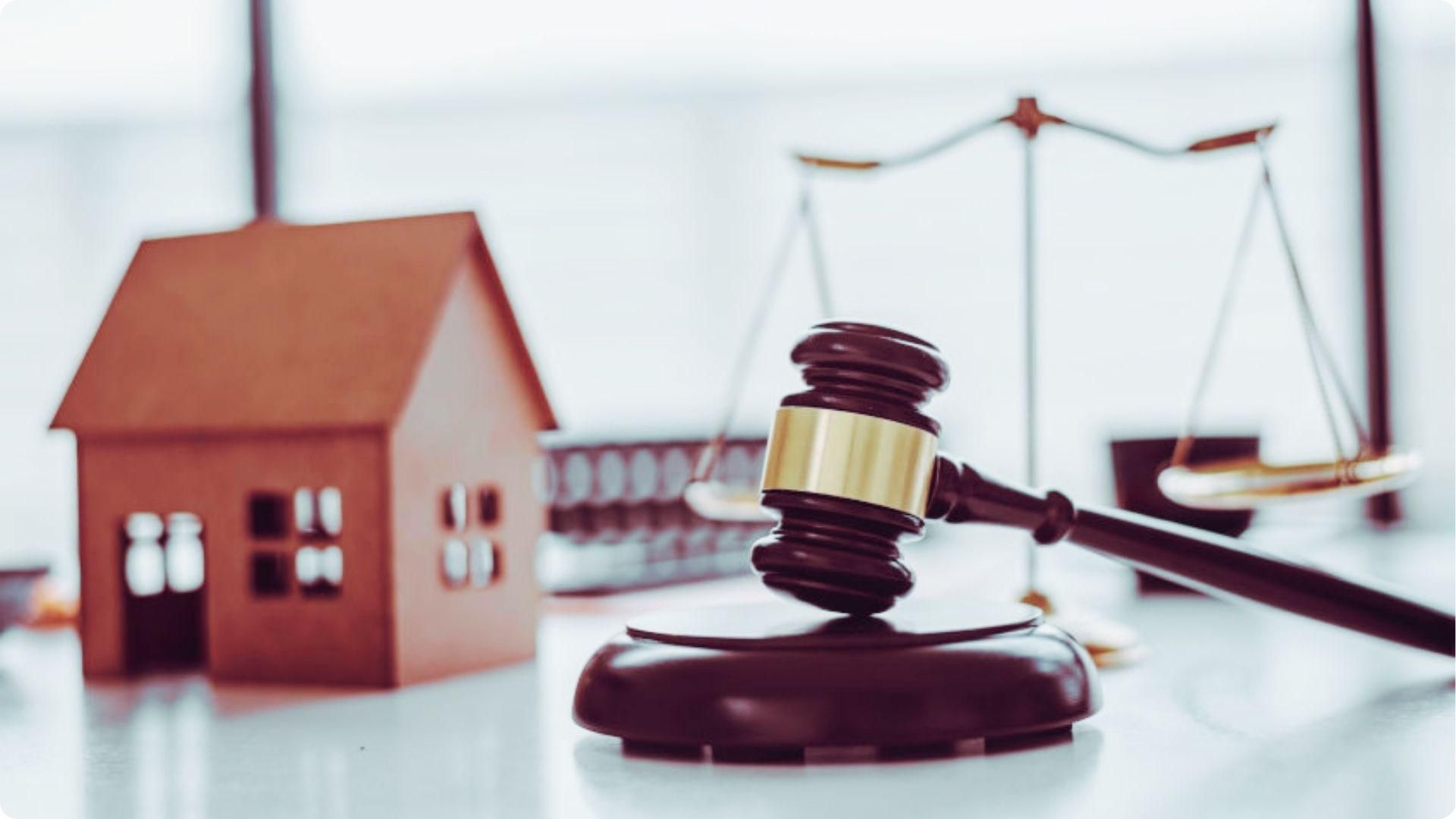 Wooden judge gavel on sounding block in focus with small brown house model and justice scales blurred in background.