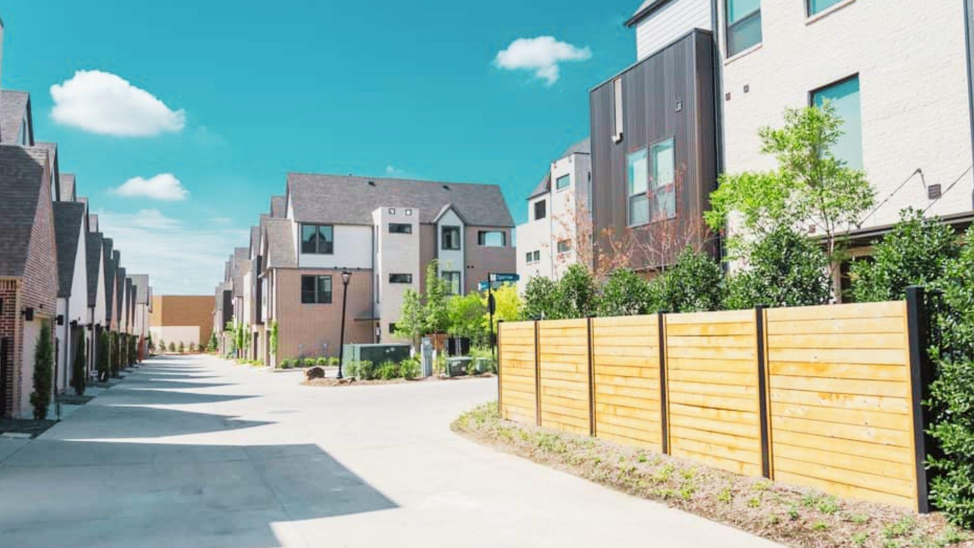 Modern townhouse-lined street with light wood fence, paved road, small trees, under bright blue sky.