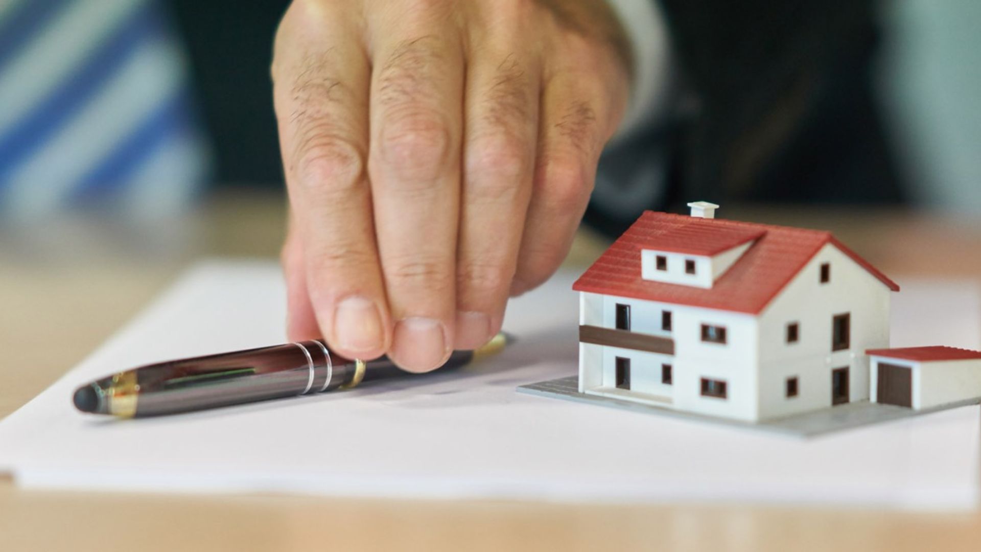 Hand reaching for pen on contract next to small white model house with red roof on desk, real estate concept.