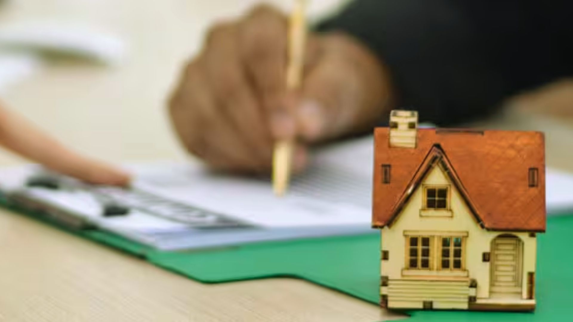 Small wooden house model on desk with person signing mortgage document in background