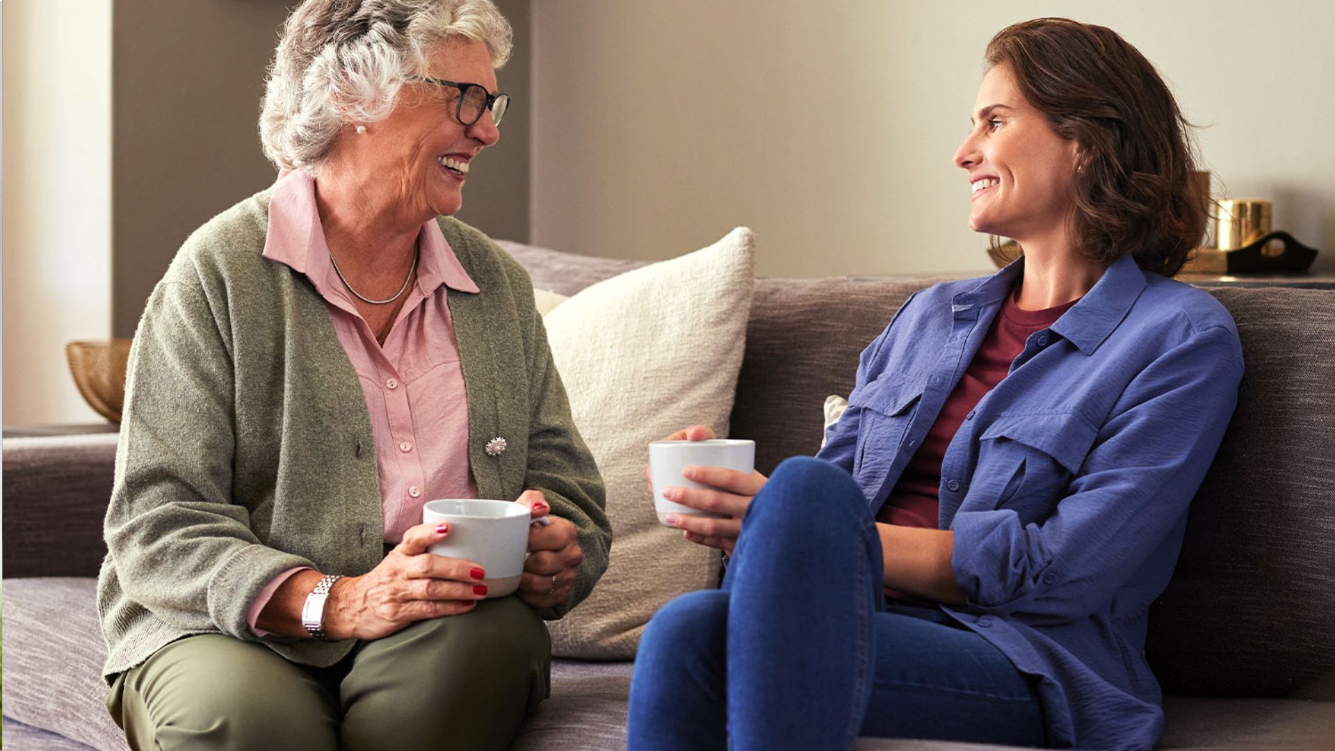Older woman with gray curly hair and glasses smiling at younger woman over coffee, both holding white mugs on gray couch.