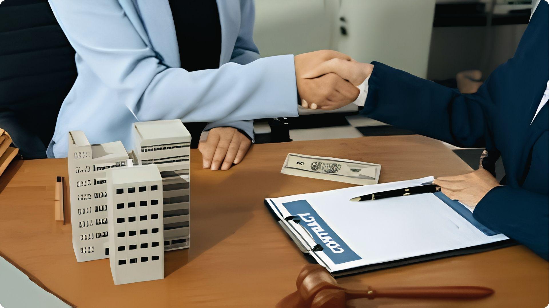 Two professionals shaking hands over building models, contract, cash and gavel finalizing commercial real estate deals.
