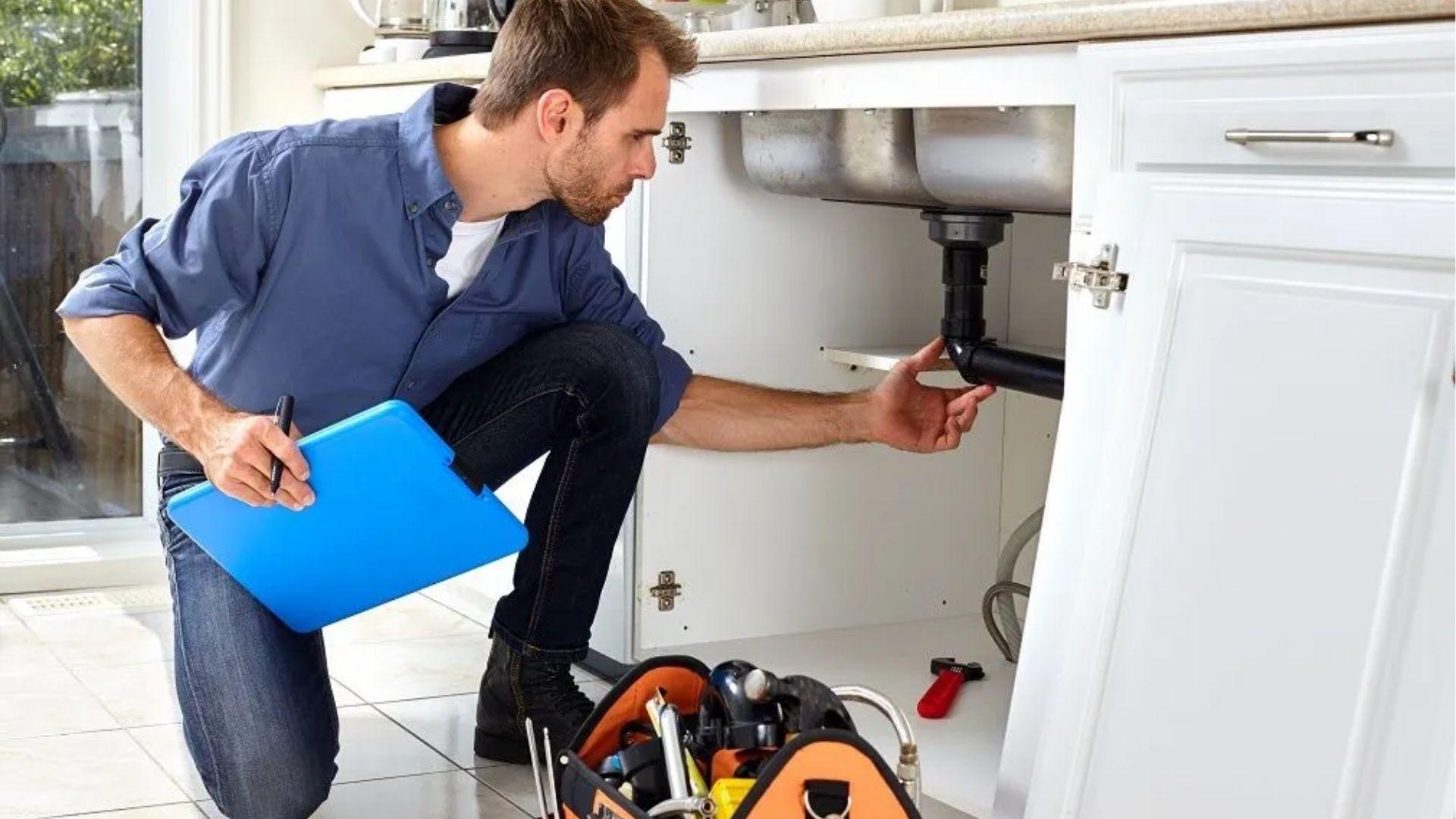 Plumber with clipboard inspecting pipes under a kitchen sink with tools