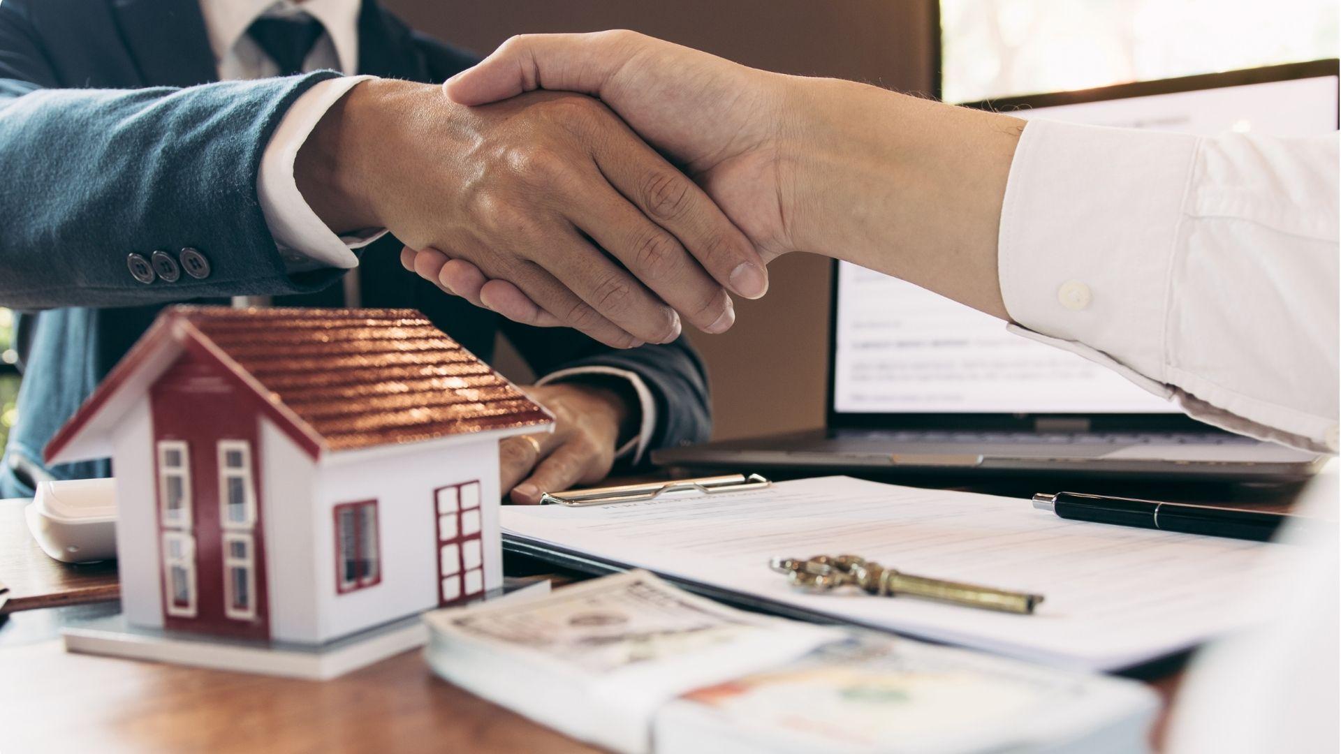Real estate agent and client shaking hands over model house, keys, and contract.