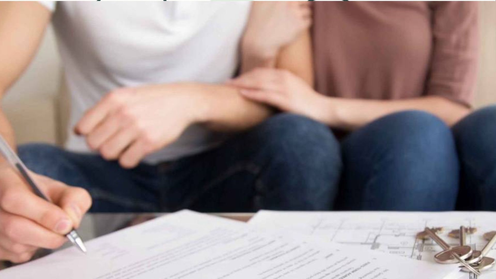 Close-up of couple on couch watching hand sign papers with house floor plans and set of keys on table foreground.