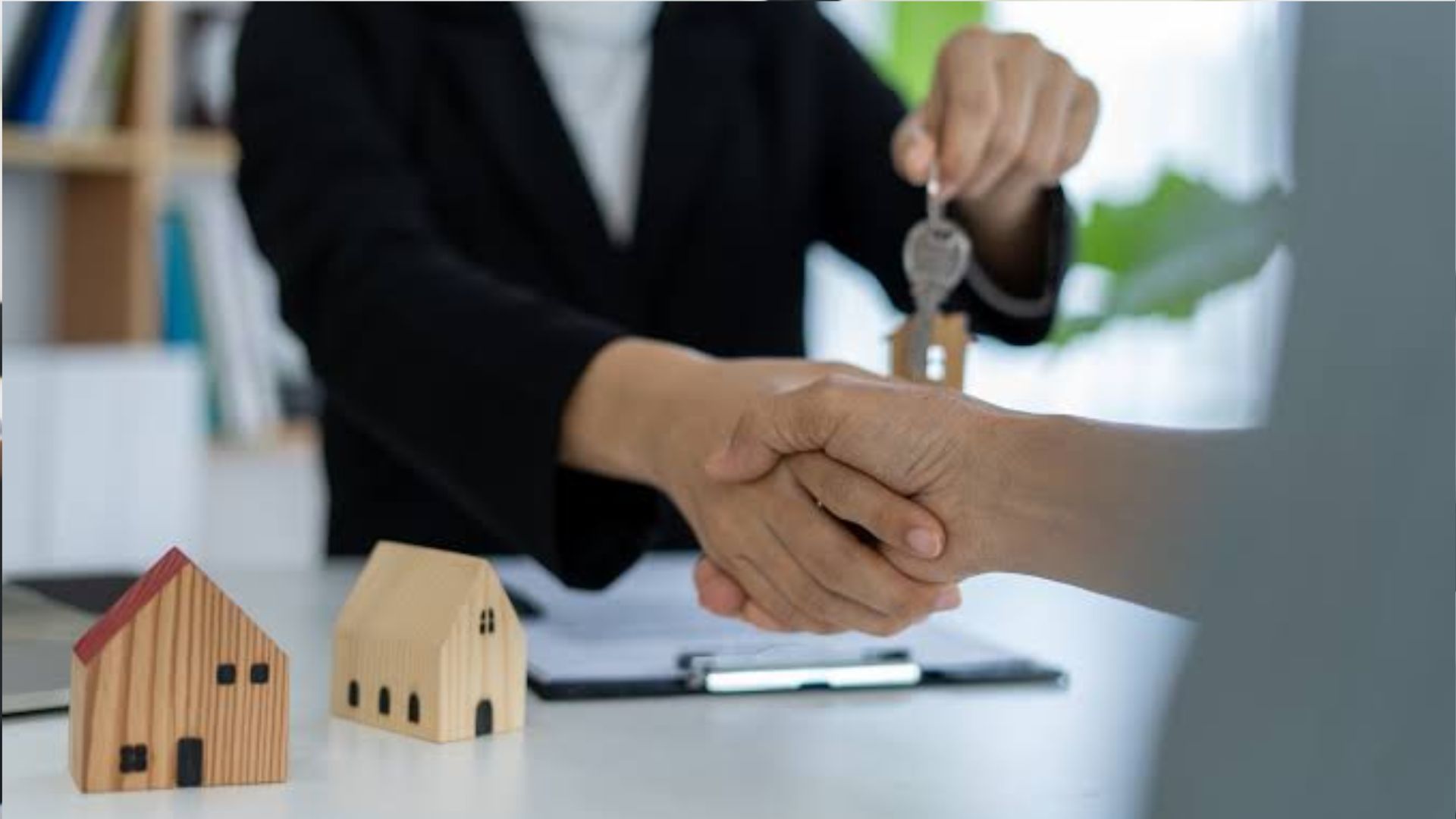Agent and client shaking hands while agent hands over a house key with a wooden house keychain, model houses on desk.