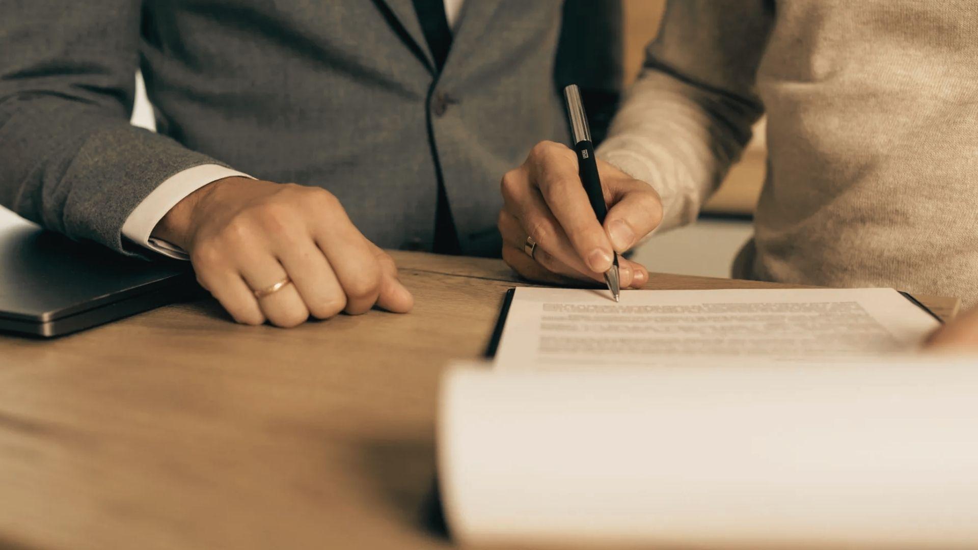 Close-up of a person signing a document on a wooden table while another person's hand rests nearby.