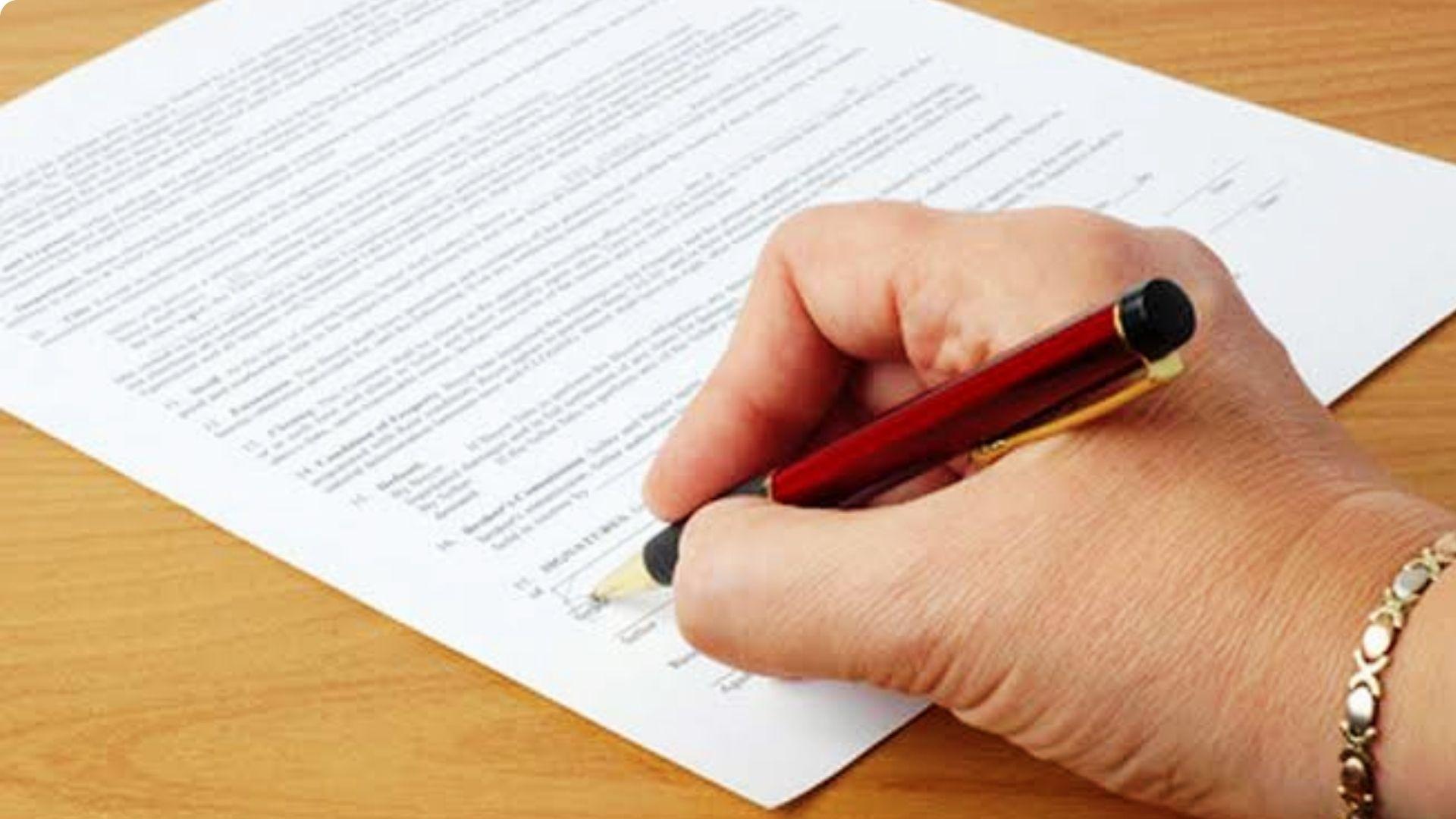 Close-up of a hand signing a warranty deed document with a red pen on a wooden table.