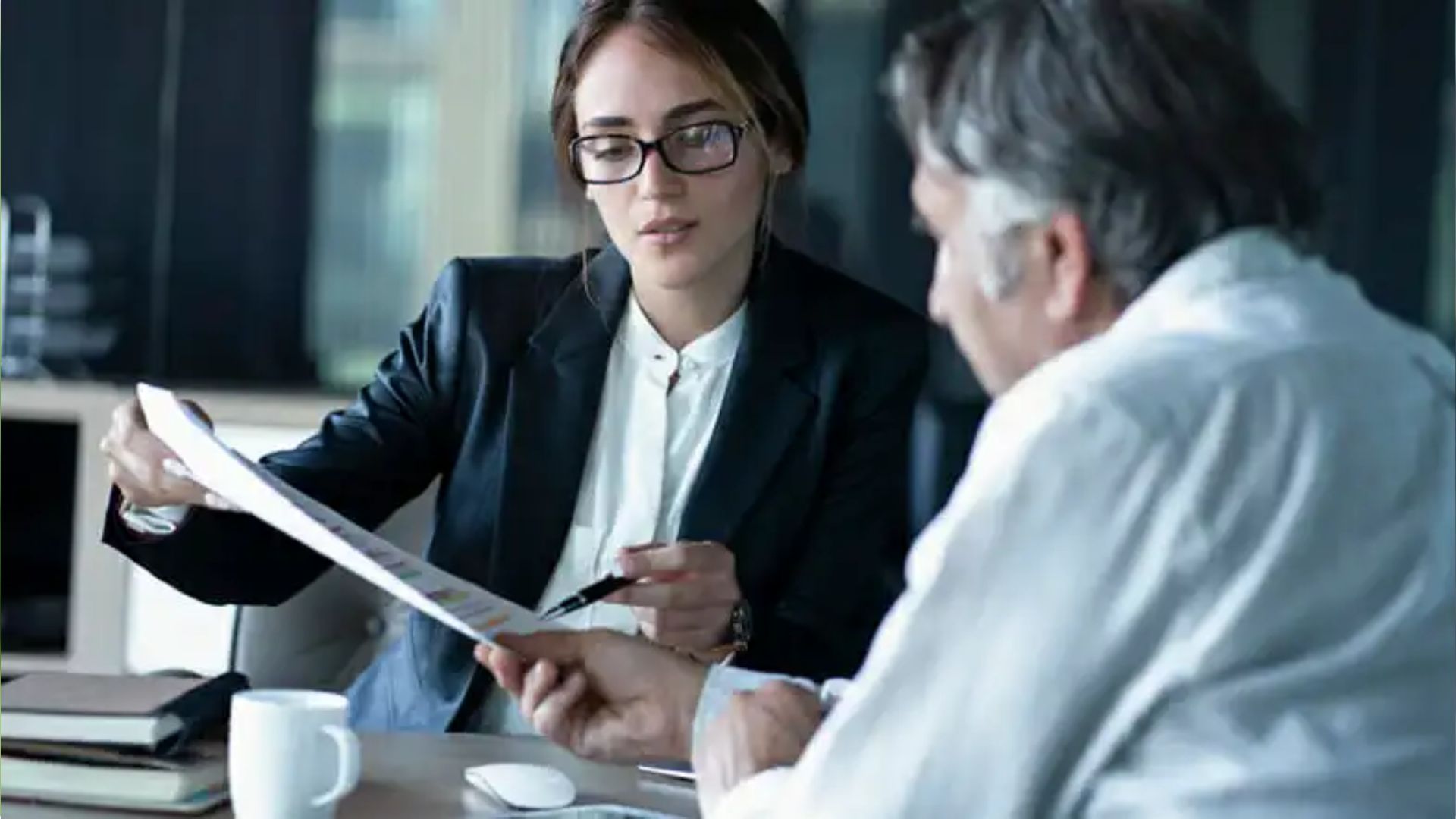 Businesswoman with glasses in black blazer showing charts document to older man at office table with books and mug.