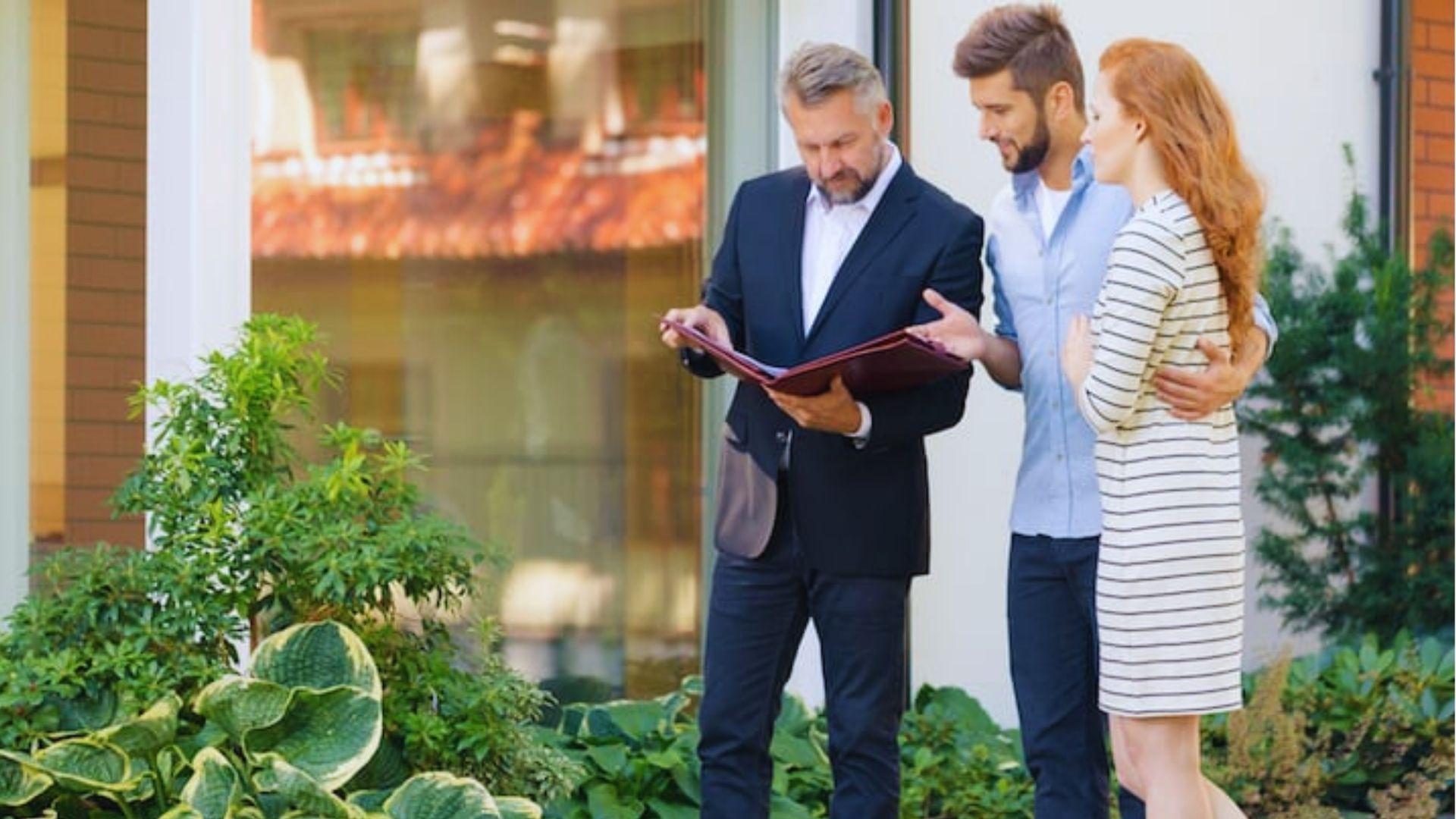 Real estate agent in suit showing documents in folder to young couple outside a house with green landscaping.