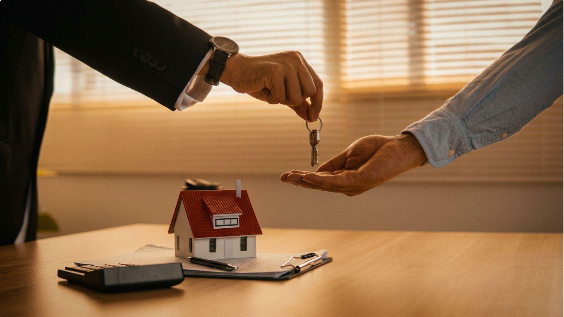 Agent handing house keys to client over model house and clipboard on desk.