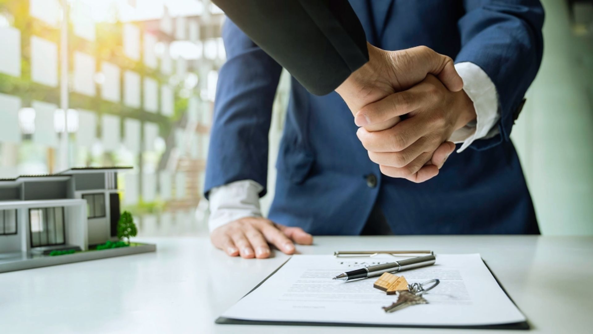 Real estate deal closed with handshake over property contract, house model and keys on office desk
