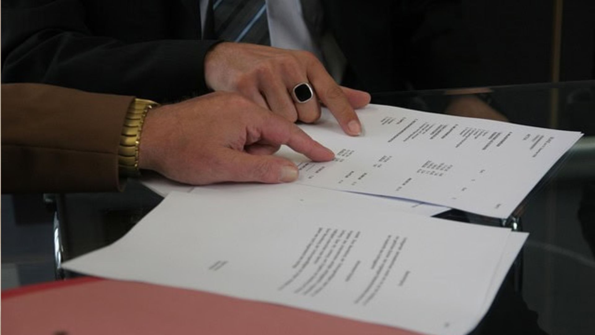 Two people pointing at contract documents on a glass table, one wearing a gold watch, other with black ring, business agreement.
