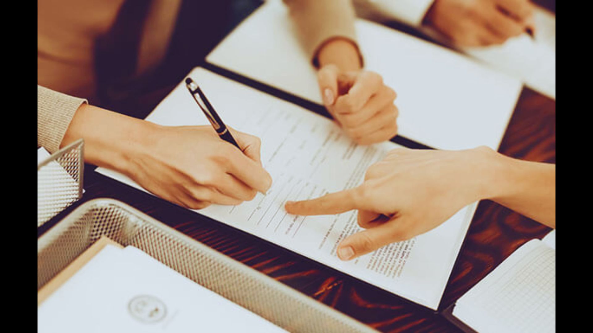 A person signing a legal document while another points to a specific section.