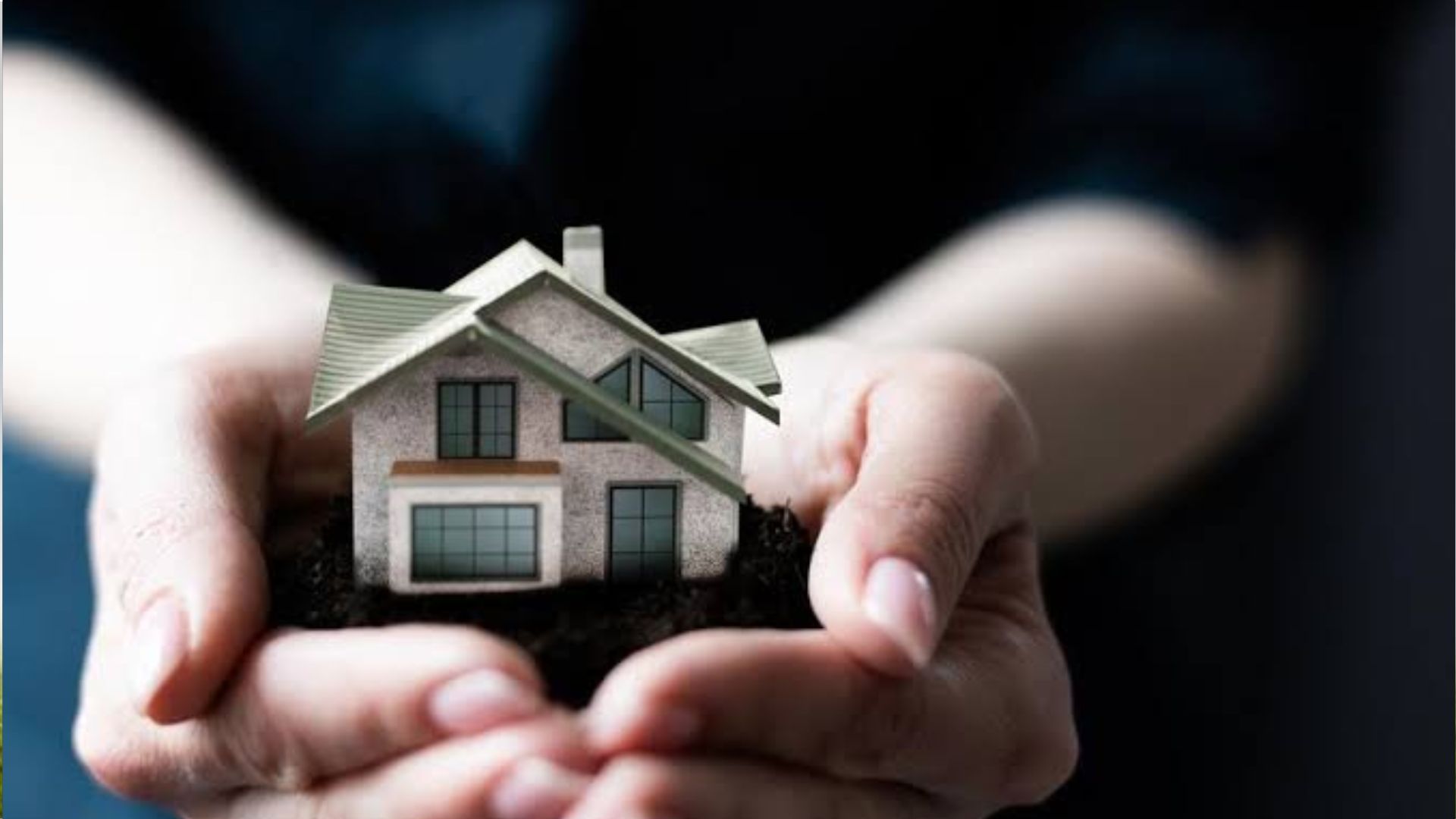 Close-up of cupped hands holding dark soil with small beige model house with green roof, symbolizing home ownership and real estate care.