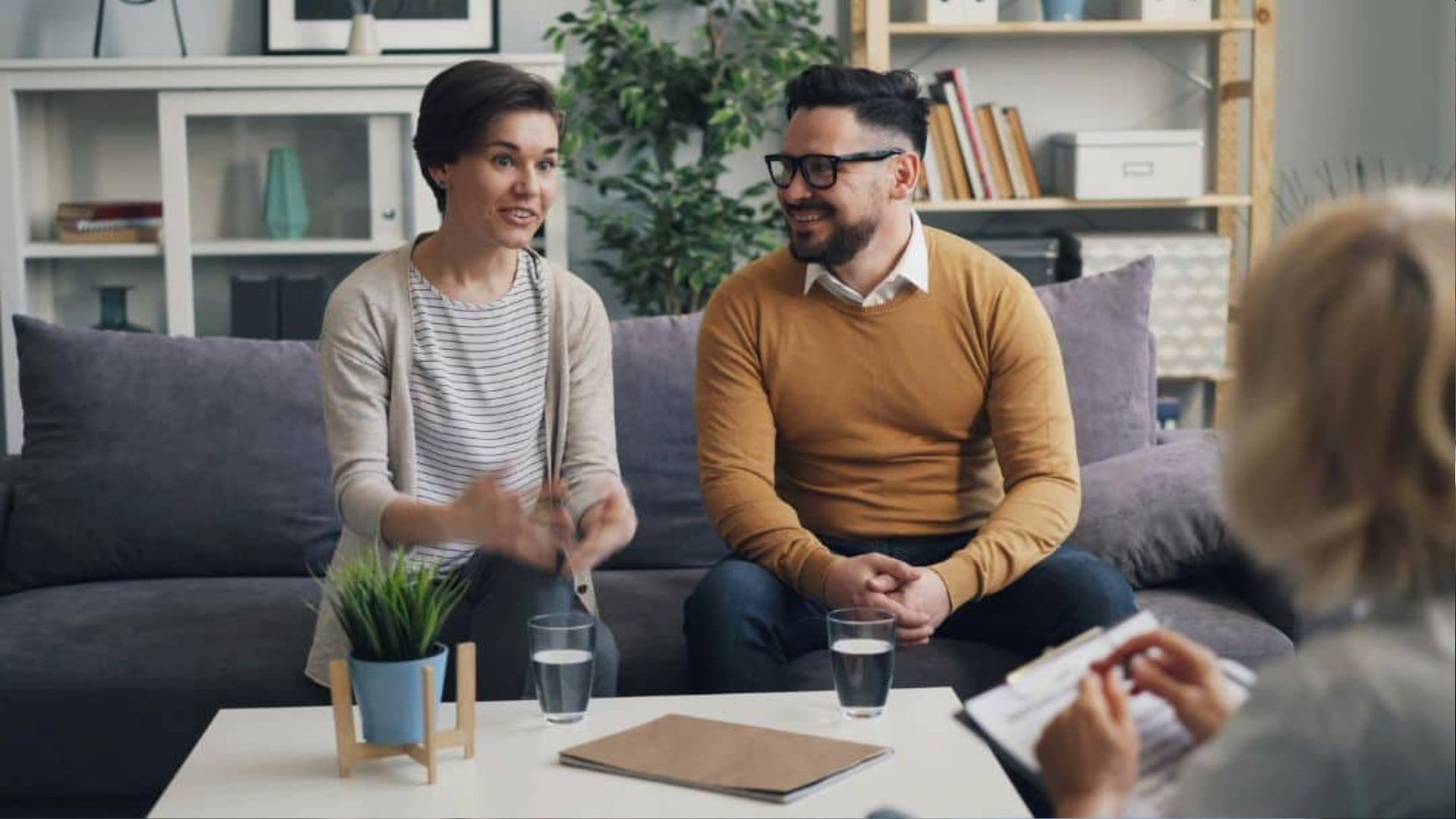 Couple sitting on gray couch talking animatedly to counselor with clipboard, coffee table with plant and water glasses.
